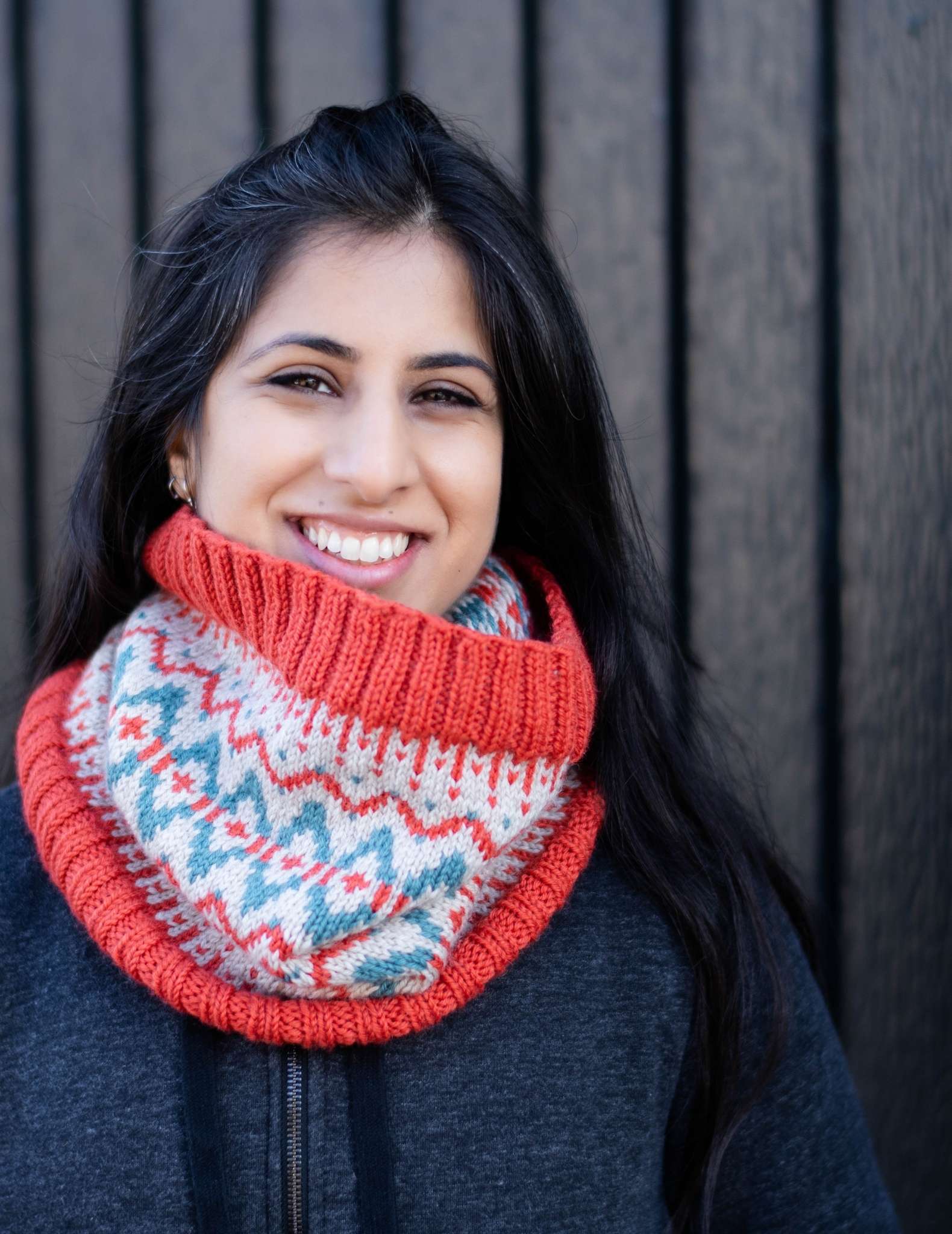 a woman with dark hair wears a red, white and teal colourwork cowl, she is smiling