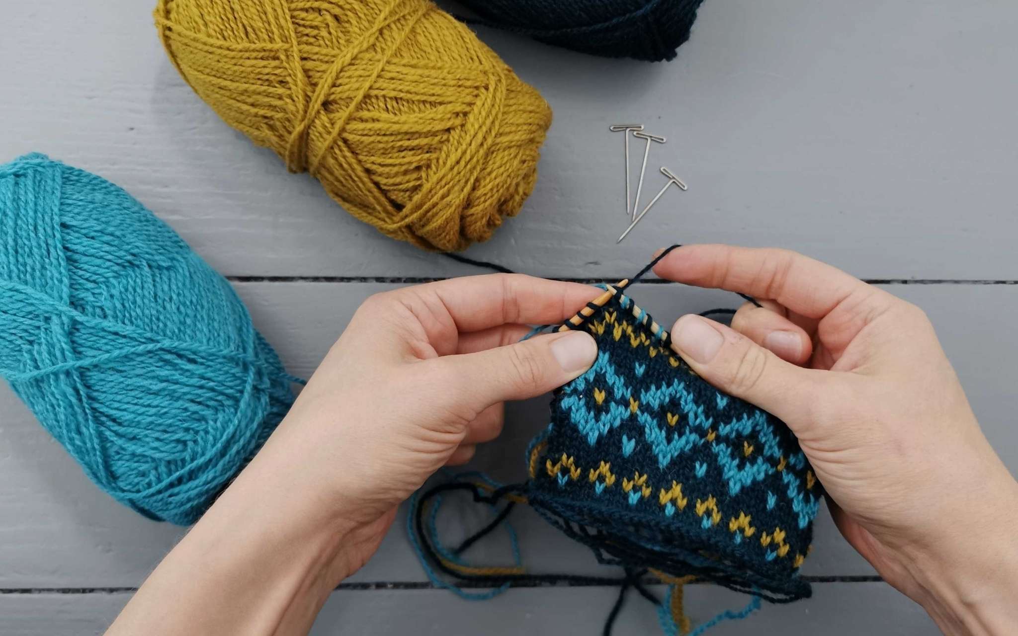 hand knitting on a swatch in three colours, yellow, blue and navy balls of yarn lie the left side
