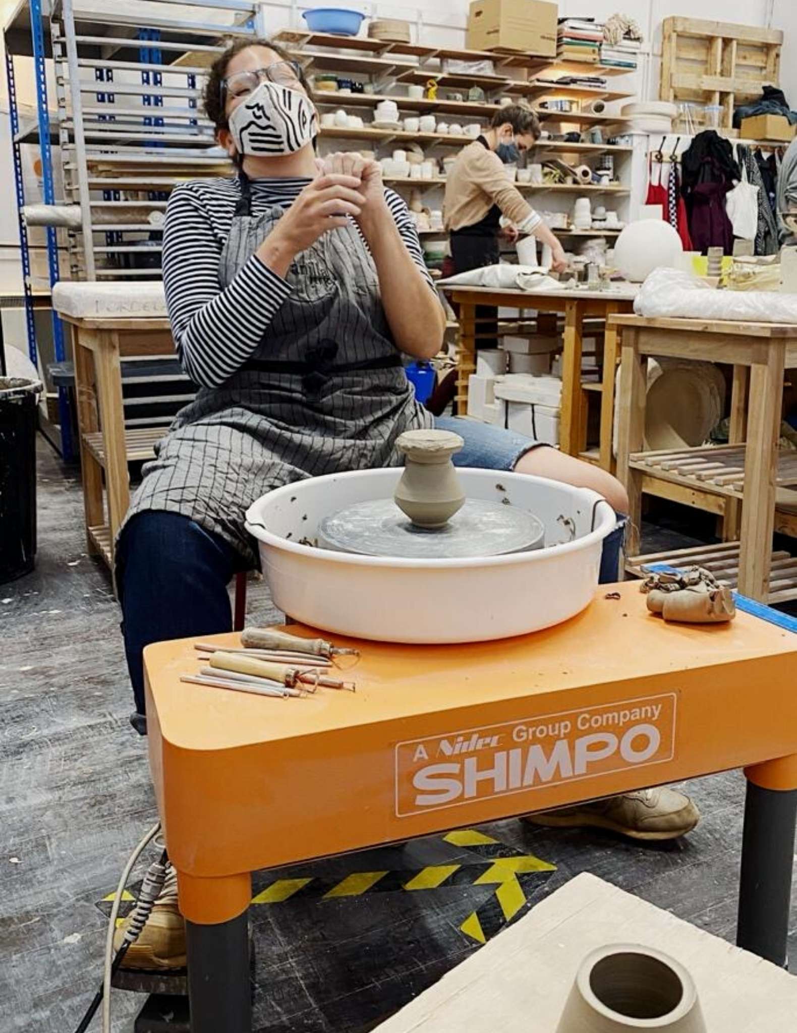 Dannie sits at a pottery wheel, wearing a face mask with her hands raised.