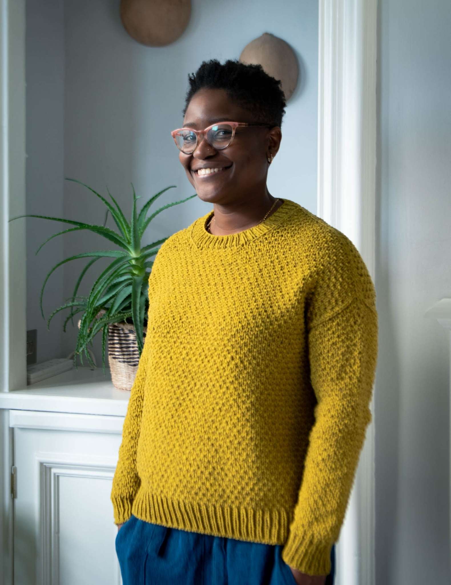 een zwarte vrouw staat binnen met een kamerplant erachter, draagt een trui met gouden textuur en heeft haar handen in haar zakken