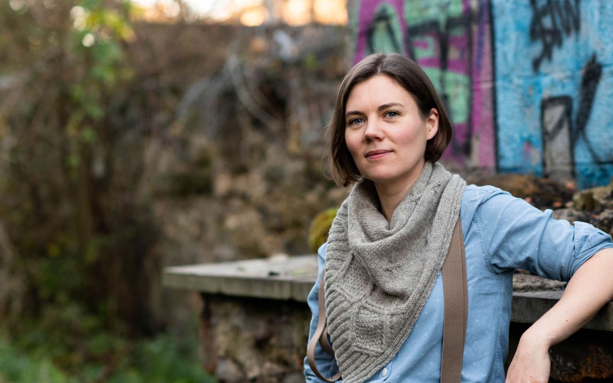 A white woman with chin length brown hair sits in front of a graffitied wall with her left arm raised and leaning on a ledge behind her. She is wearing a neutral cabled shawl around her neck, with a blue shirt and brown braces visible underneath.