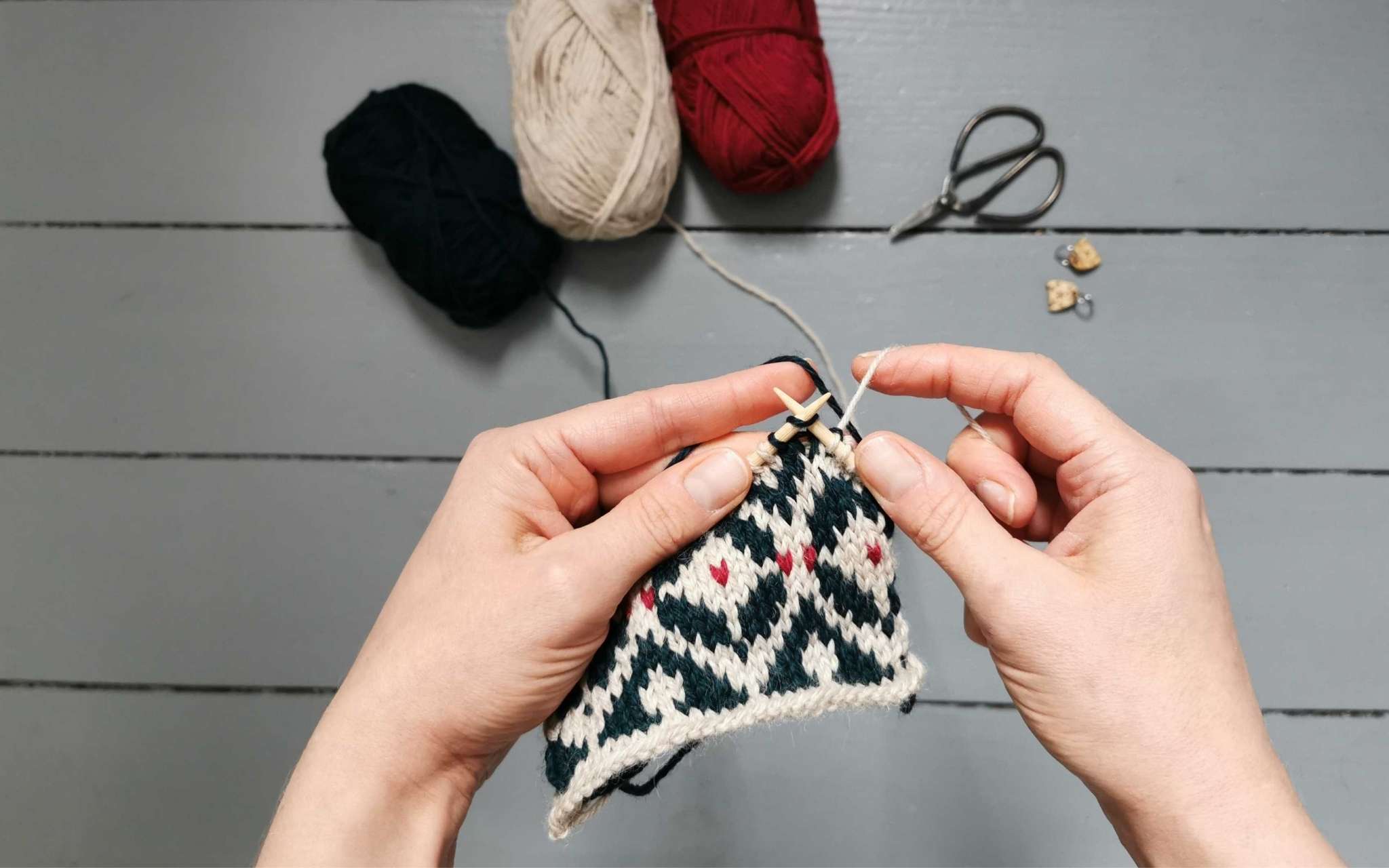 two hands hold a piece of colourwork knitting on the needles, in cream, dark green and dark pink.