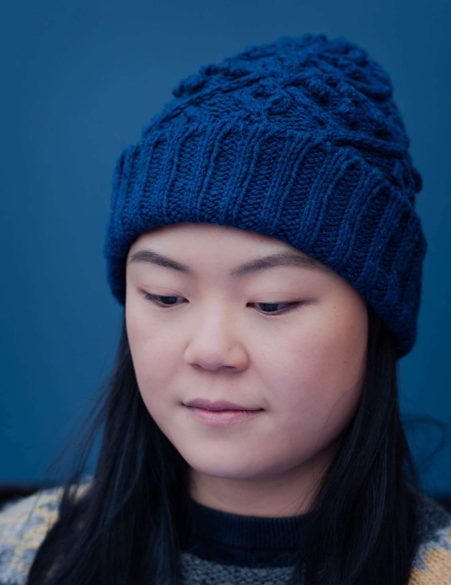 a close up of a young woman with straight dark hair looking down, she is wearing a cabled blue hat against a blue background