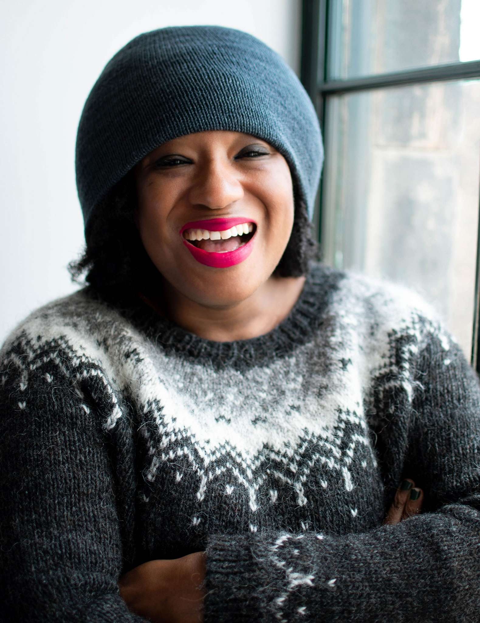 A close up of a black woman with bright pink lipstick wearing a teal beanie hat and colourwork sweater in blacks and neutral colours.