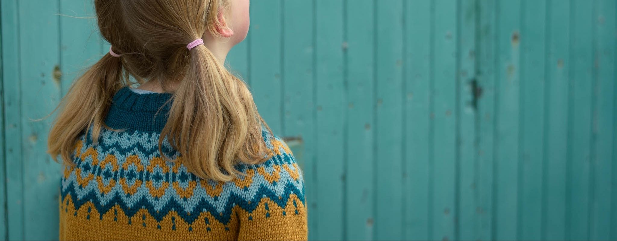 Cropped image showing a blond child with pigtails looking away from the camera, wearing the Brunstane Sweater in the vintage palette. The background is a turquoise painted wooden wall.
