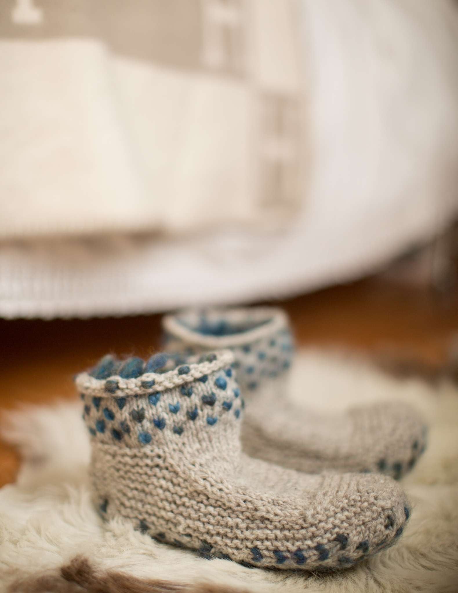 a pair of pale knitted slippers with a teal dot pattern around the cuff sit next to each other on a cream rug