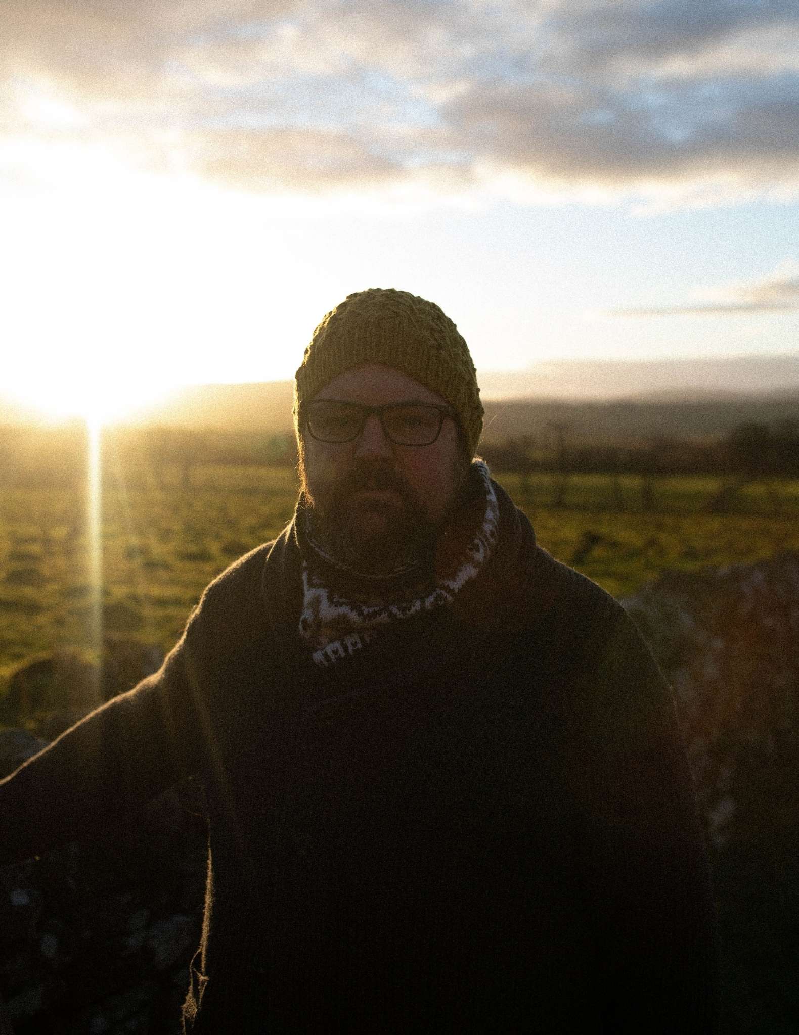 an under exposed, and dark photo of a man with a beard and glasses standing in a field with the sun behind. The photo is so dark that few details are visible.