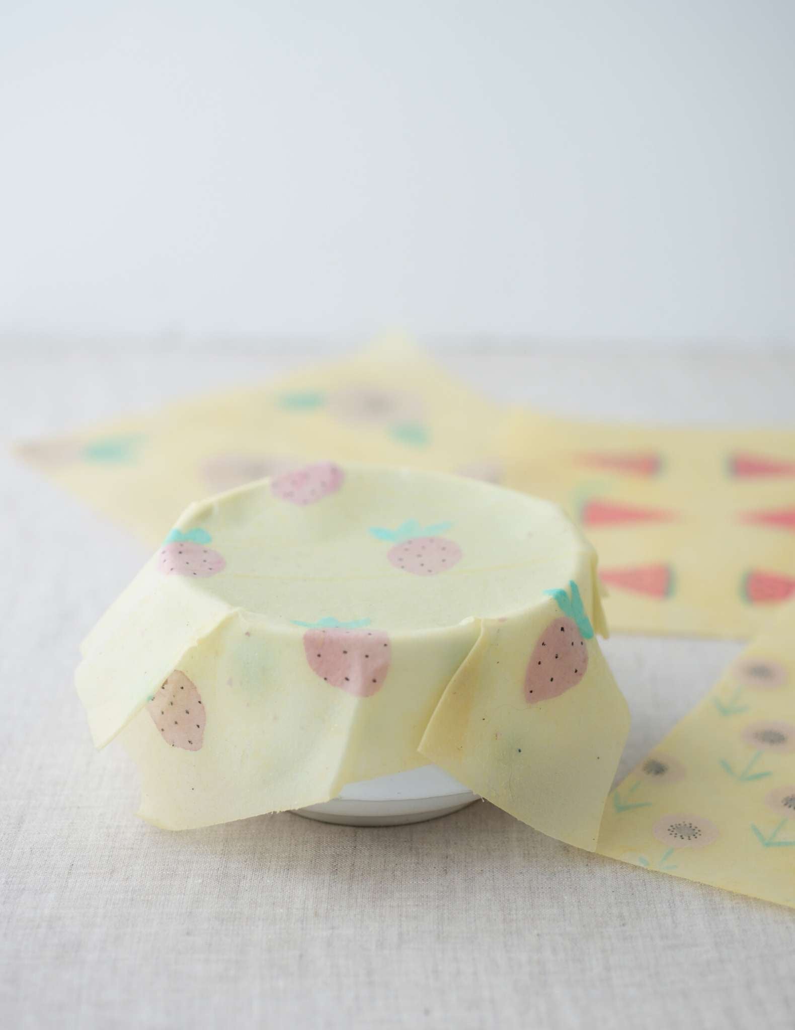 a white bowl covered in a pale food wrap that clings to the top edge of the bowl. The food wrap is handprinted with a strawberry motif and other, unused wraps lay behind.