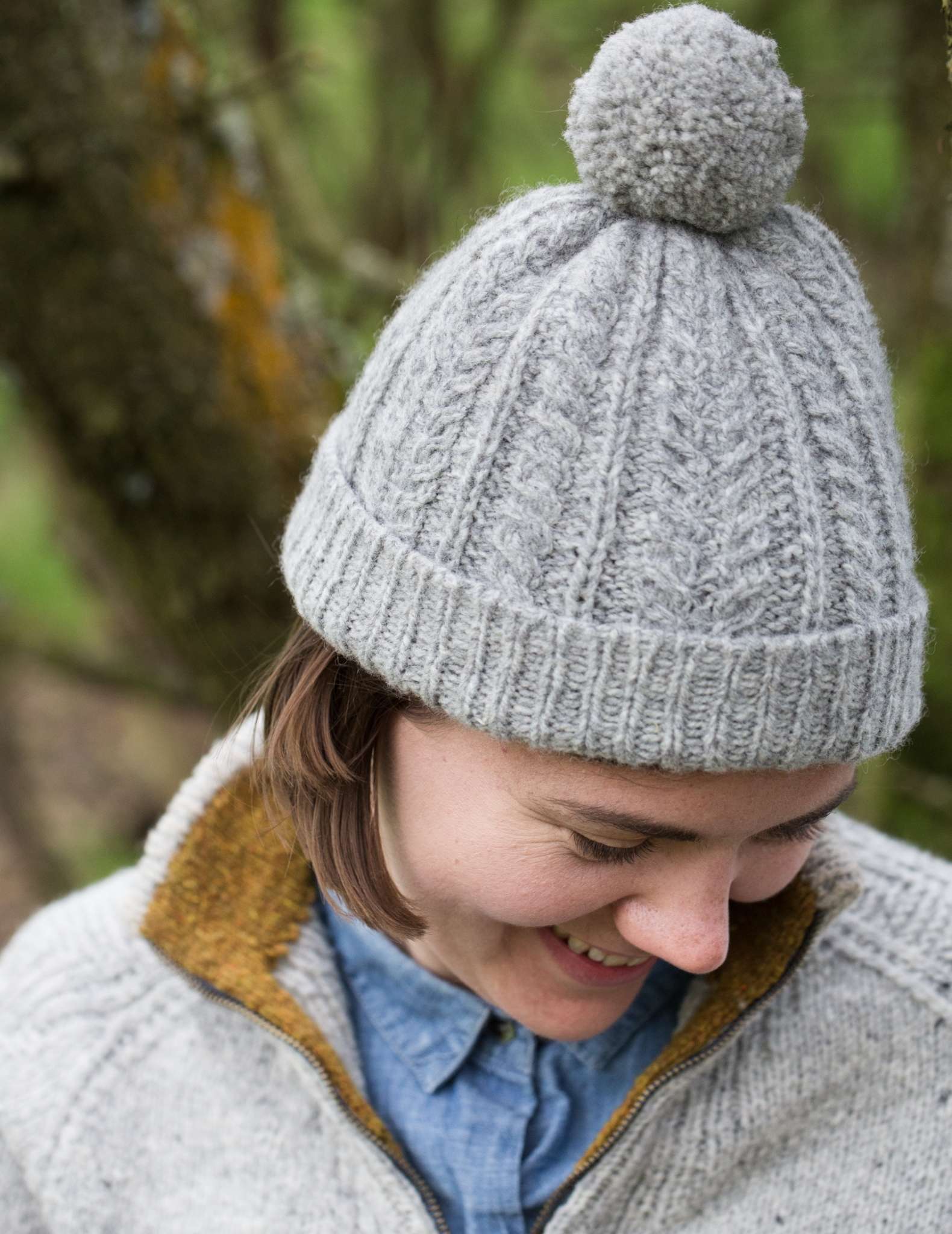 a woman with brown hair looks down and is laughing, she wears a pale grey cabled hat with a pom pom.