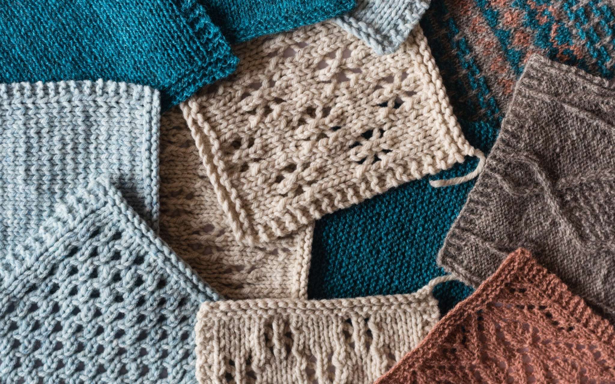 A pile of swatches, all laying flat and overlapping so that they are all that is visible. They are knitted in lace and textured stitch patterns in shades of beige, blues and brown.s