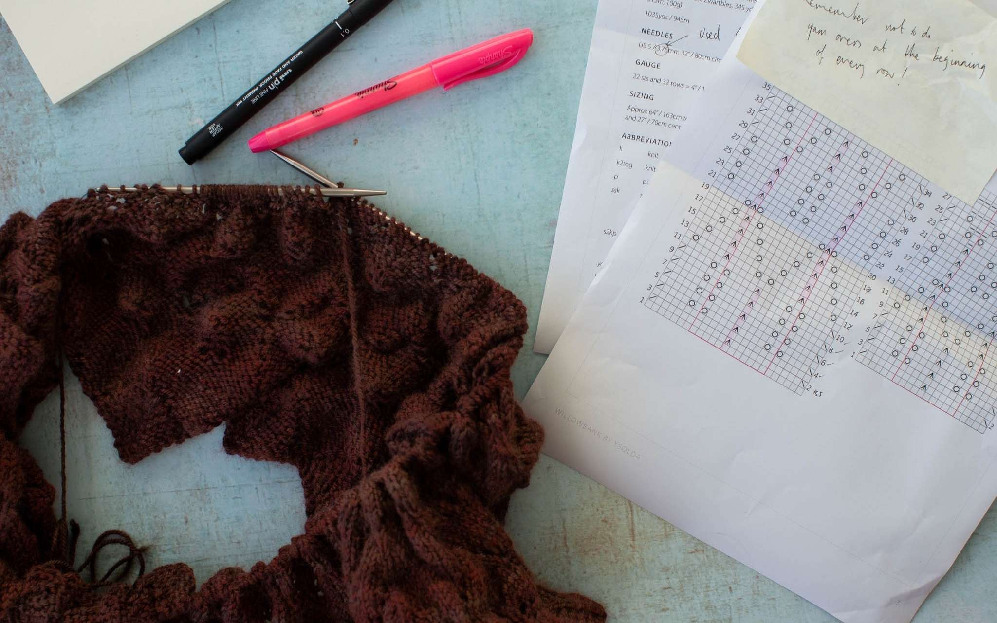 A brown lace knitting project lies on a blue surface next to a black and pink pen, the sheets of a paper knitting pattern and post it notes.