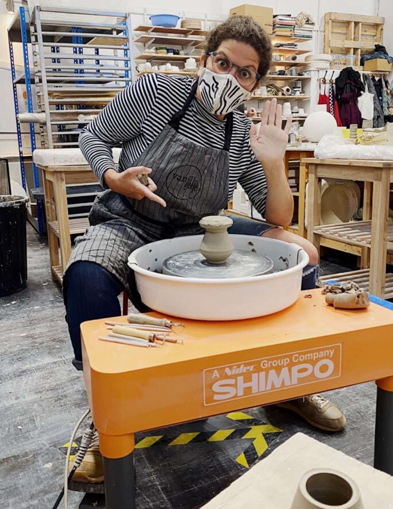 Danni leans over a pottery wheel, wearing a face mask and points at the clay she is forming.