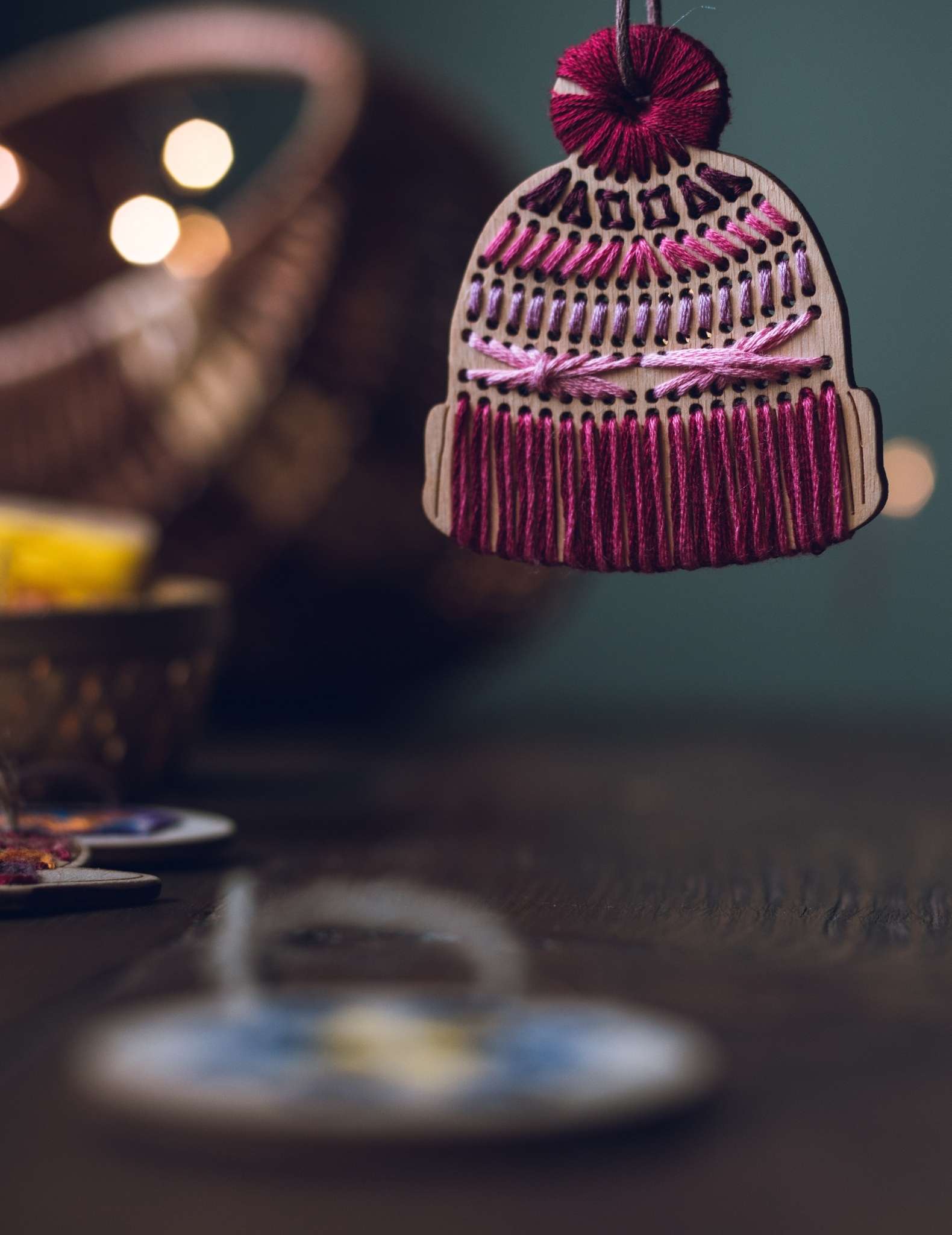 A close up of a wooden ornament shaped like a knitted hat with pom pom, stitched with pink threads.