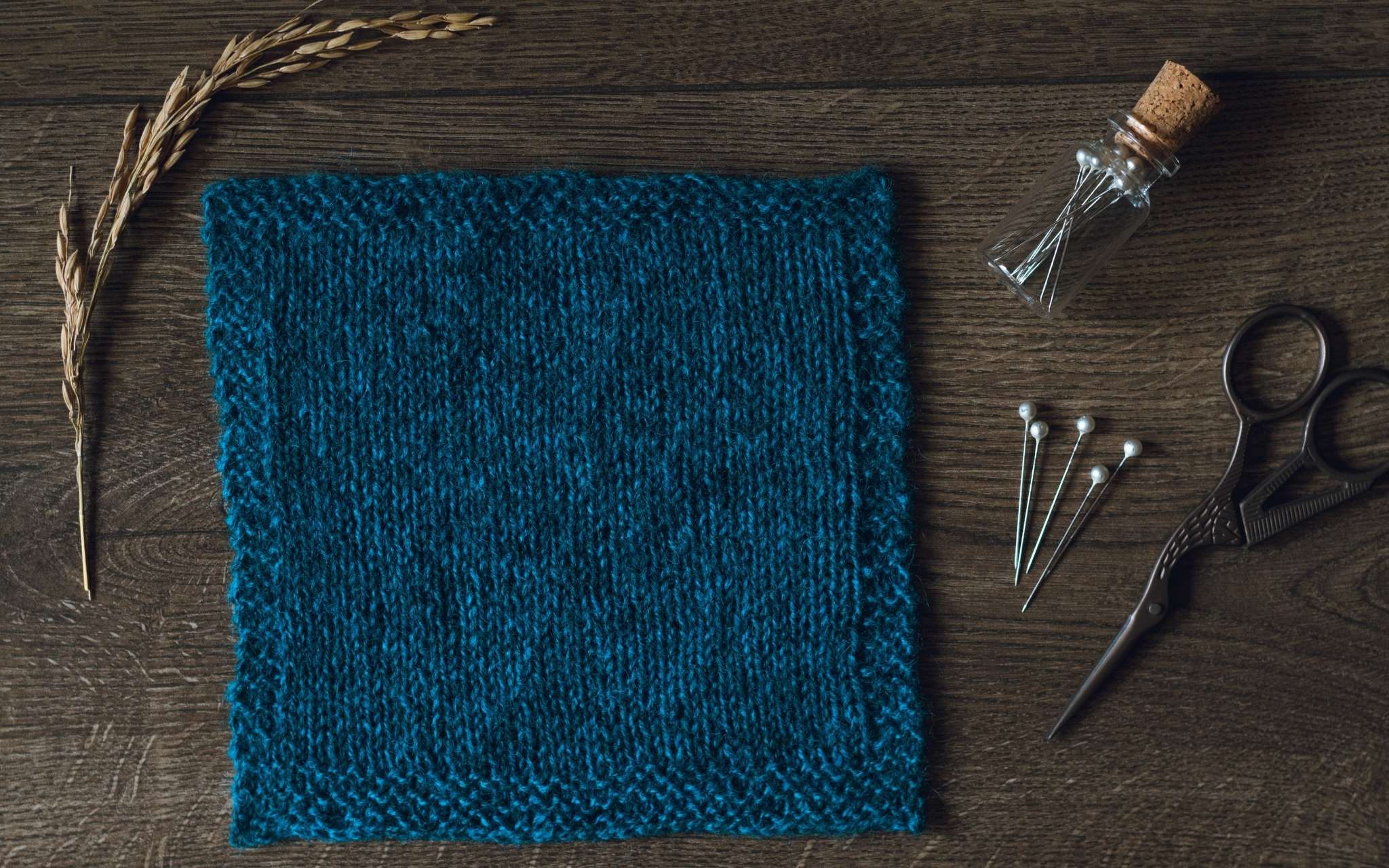 A swatch of stocking stitch in dark blue laid flat on a wooden surface. There are some dried stems, scissors and a bottle of pints to the side.
