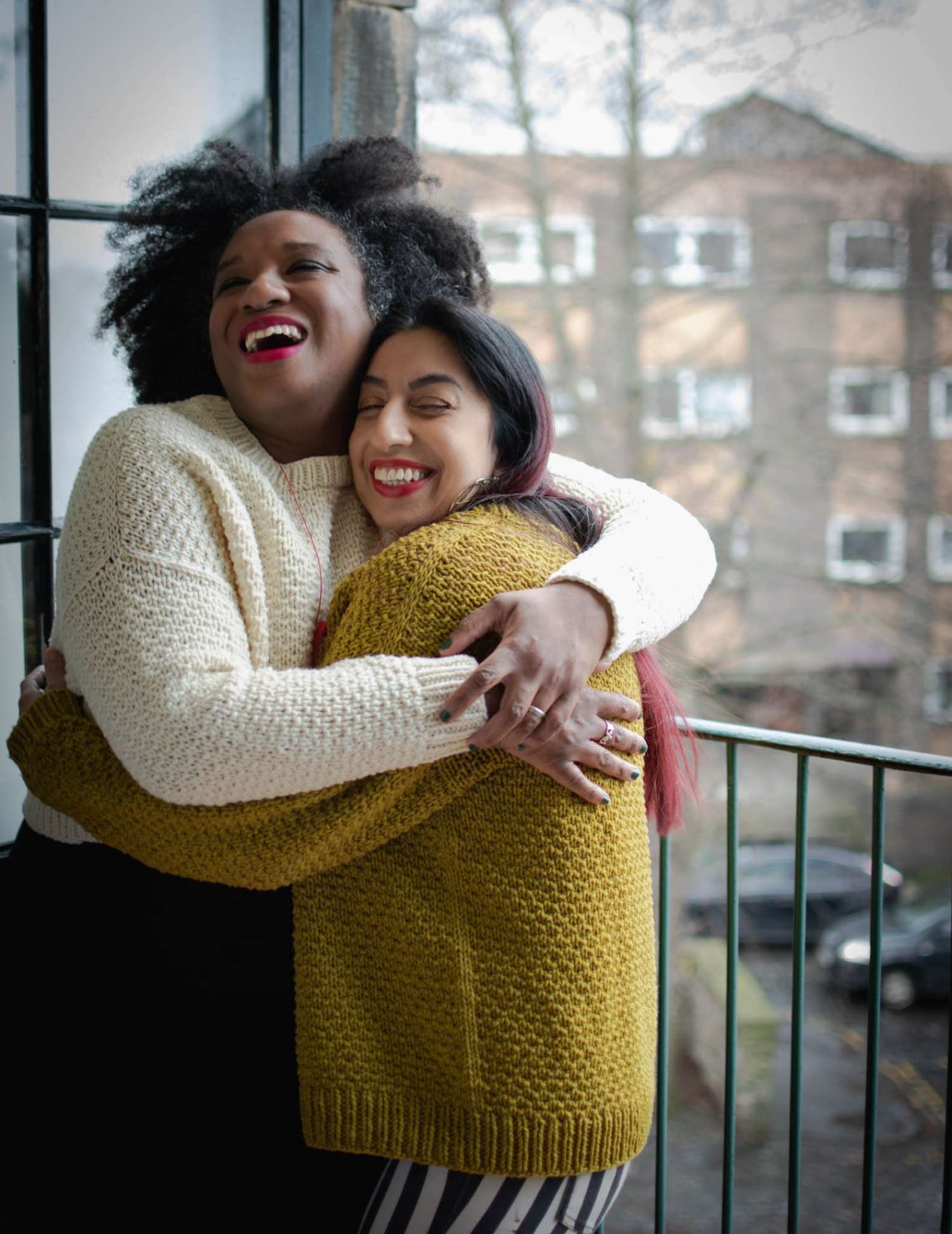 Two models stand indoors in front of a window, looking out onto a street. One wears a cream sweater and the other wears a gold sweater, they have their arms around each other in a tight hug and are grinning.