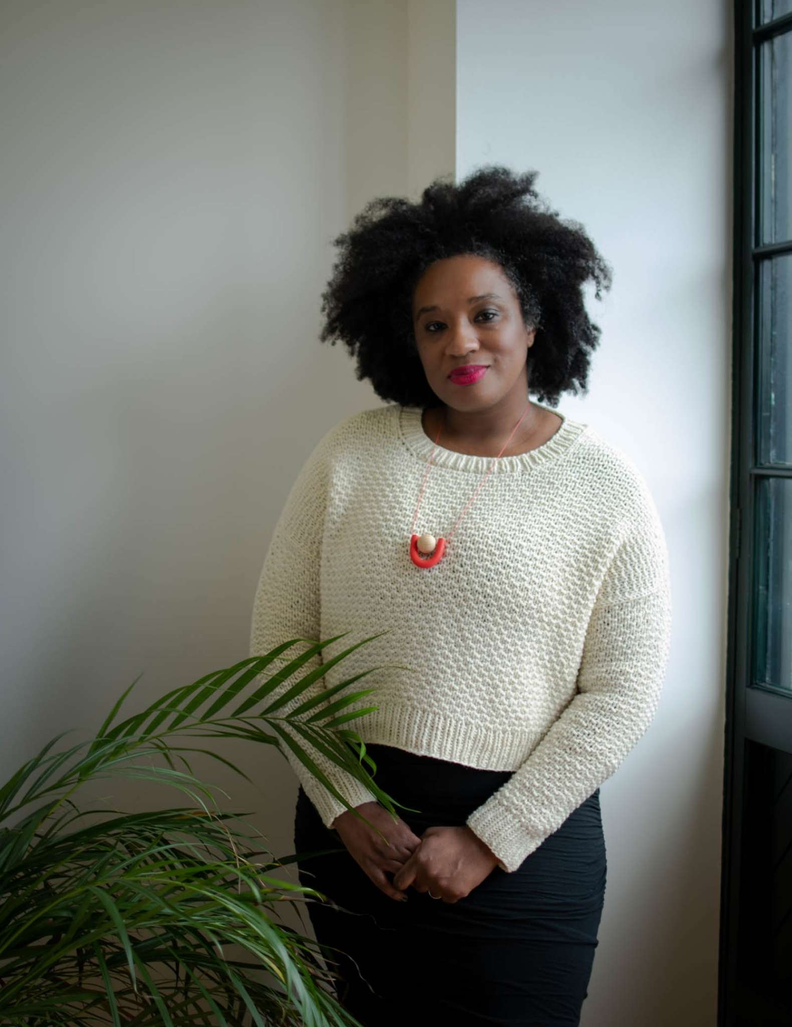 een zwarte vrouw met een korte witte trui staat naast een kamerplant, haar handen zijn voor haar samengeklemd en ze draagt een oranje ketting