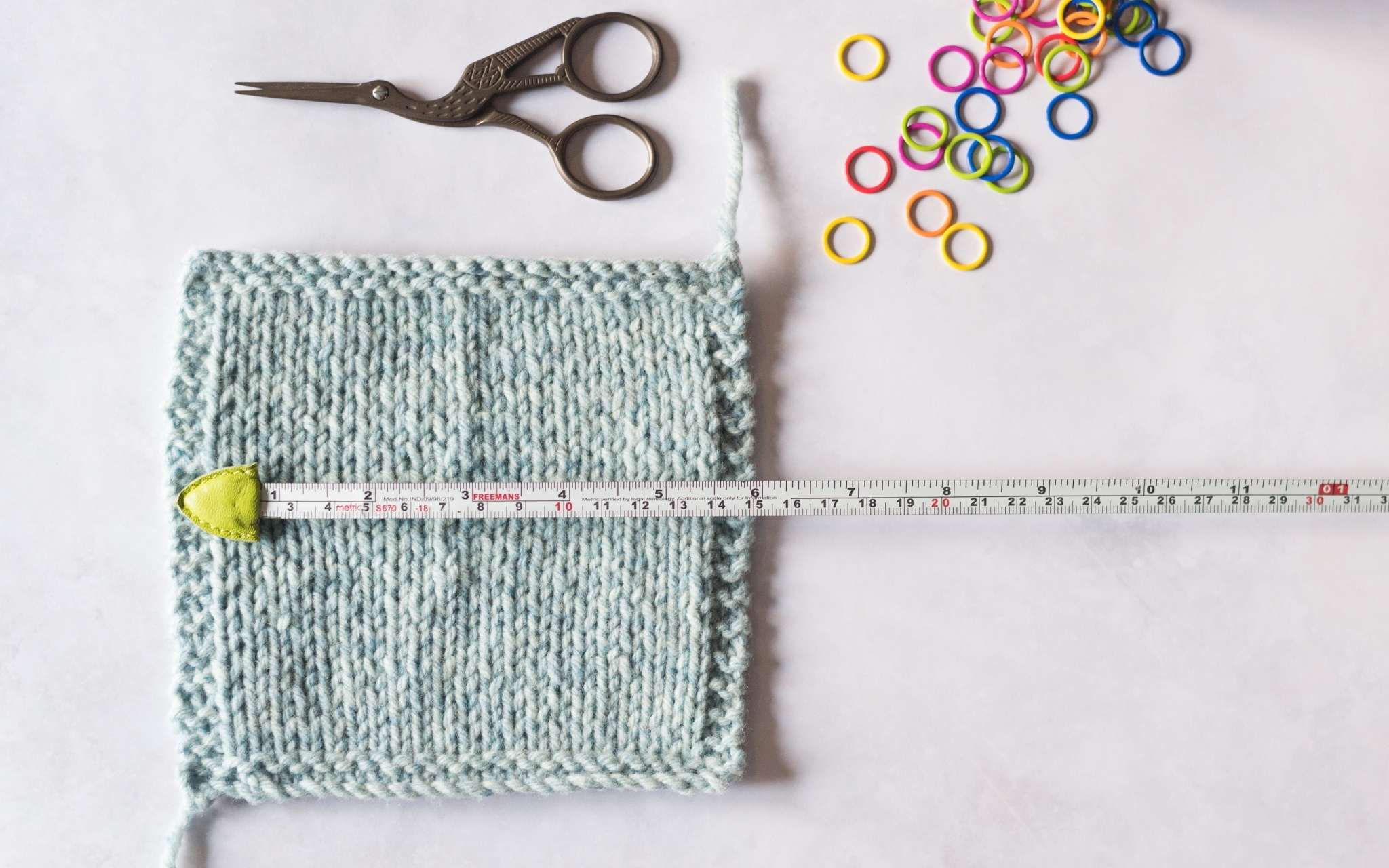 A pale blue stocking stitch swatch lies flat with a measuring tape extended across the centre. At the top of the image is a pair of small scissors and some brightly coloured stitch markers.
