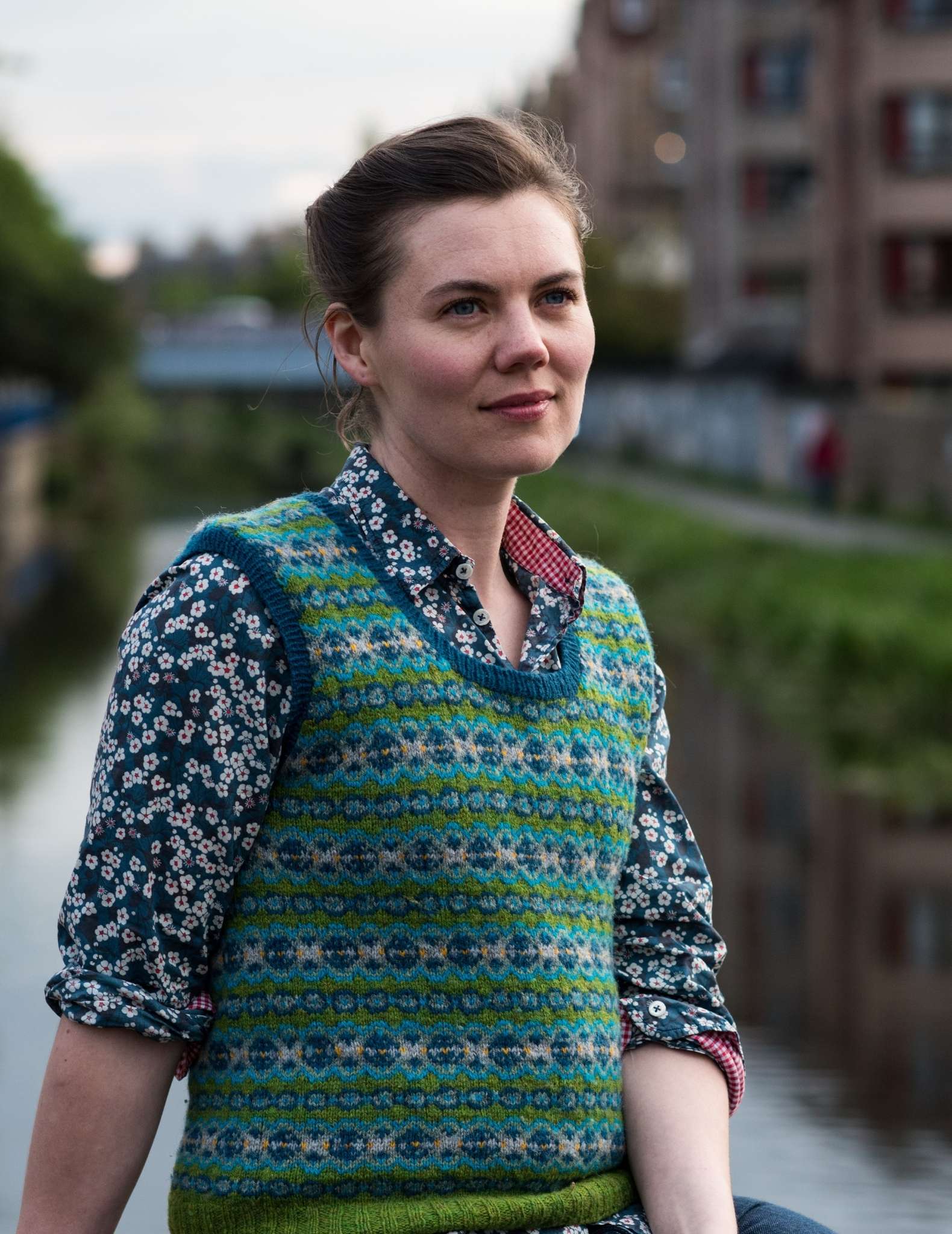 a young white woman wears a colourwork vest in greens and blues over a patterned blue shirt. Her hair is tied back and behind is the water of a canal and buildings to the side.