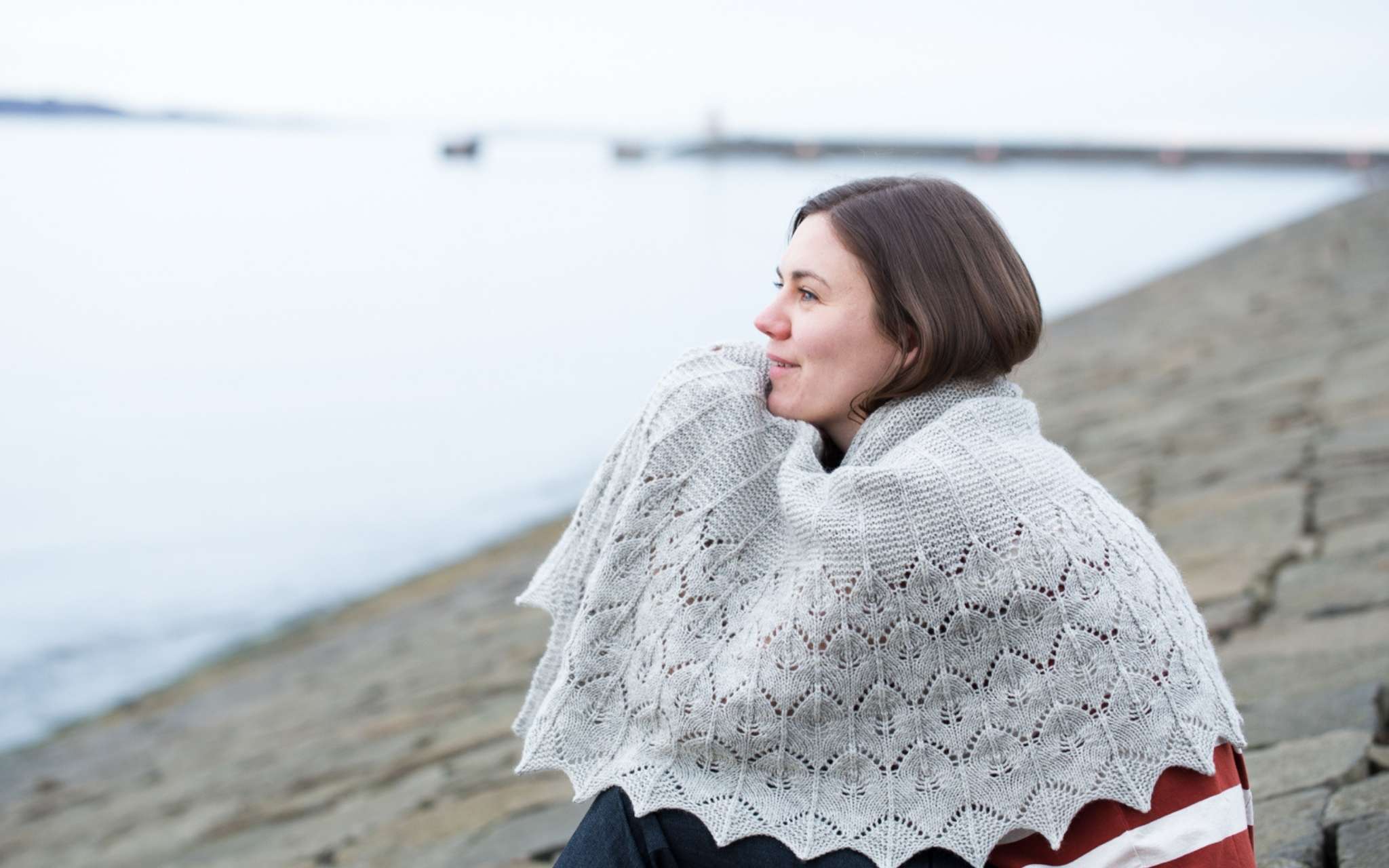 A white woman with brown hair sits huddled on a beach, looking out to sea. She has a large grey shawl wrapped around her and is resting her head in her hands, which are underneath the shawl.