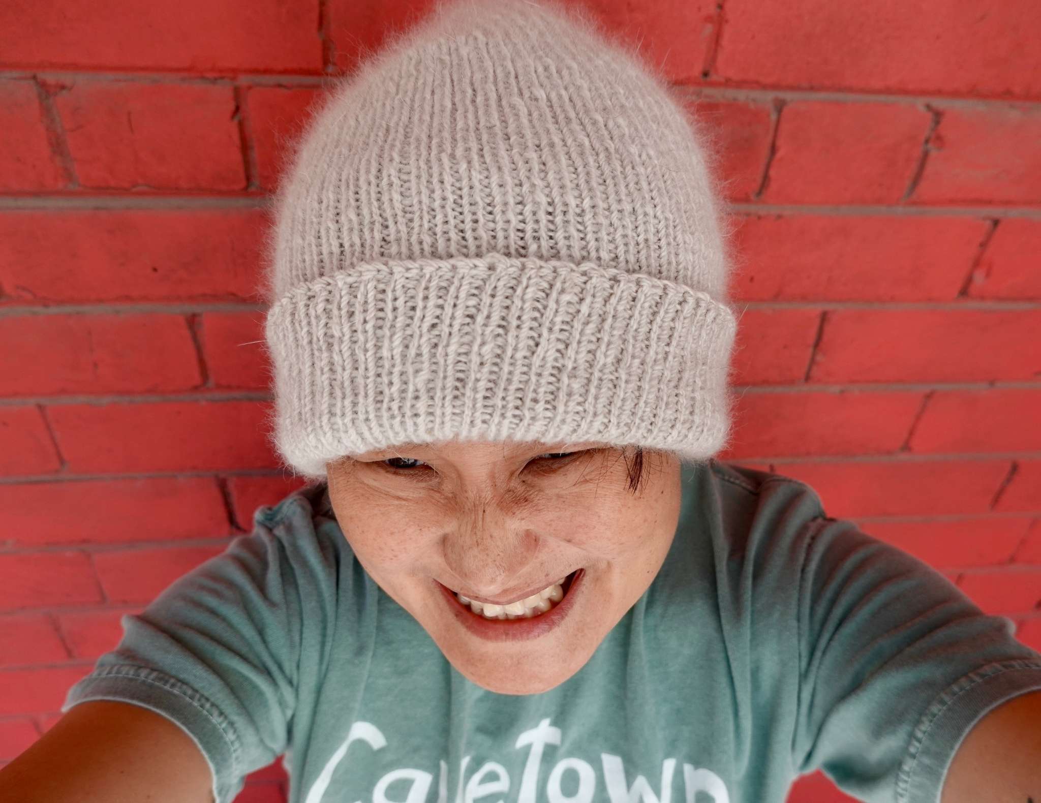 Selfie of a grinning person with freckles and short black hair wearing a fluffy grey ribbed hat. They're holding the camera and leaning on a red brick wall. 