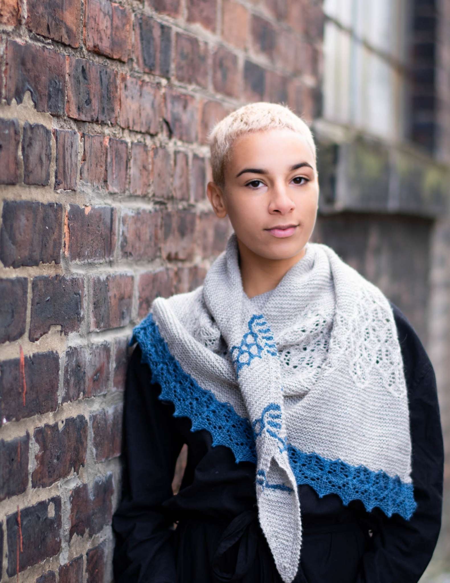 A person of colour with very short pale hair stands with against a brick wall. They wear a grey shawl with blue contrast edging and a black jacket.