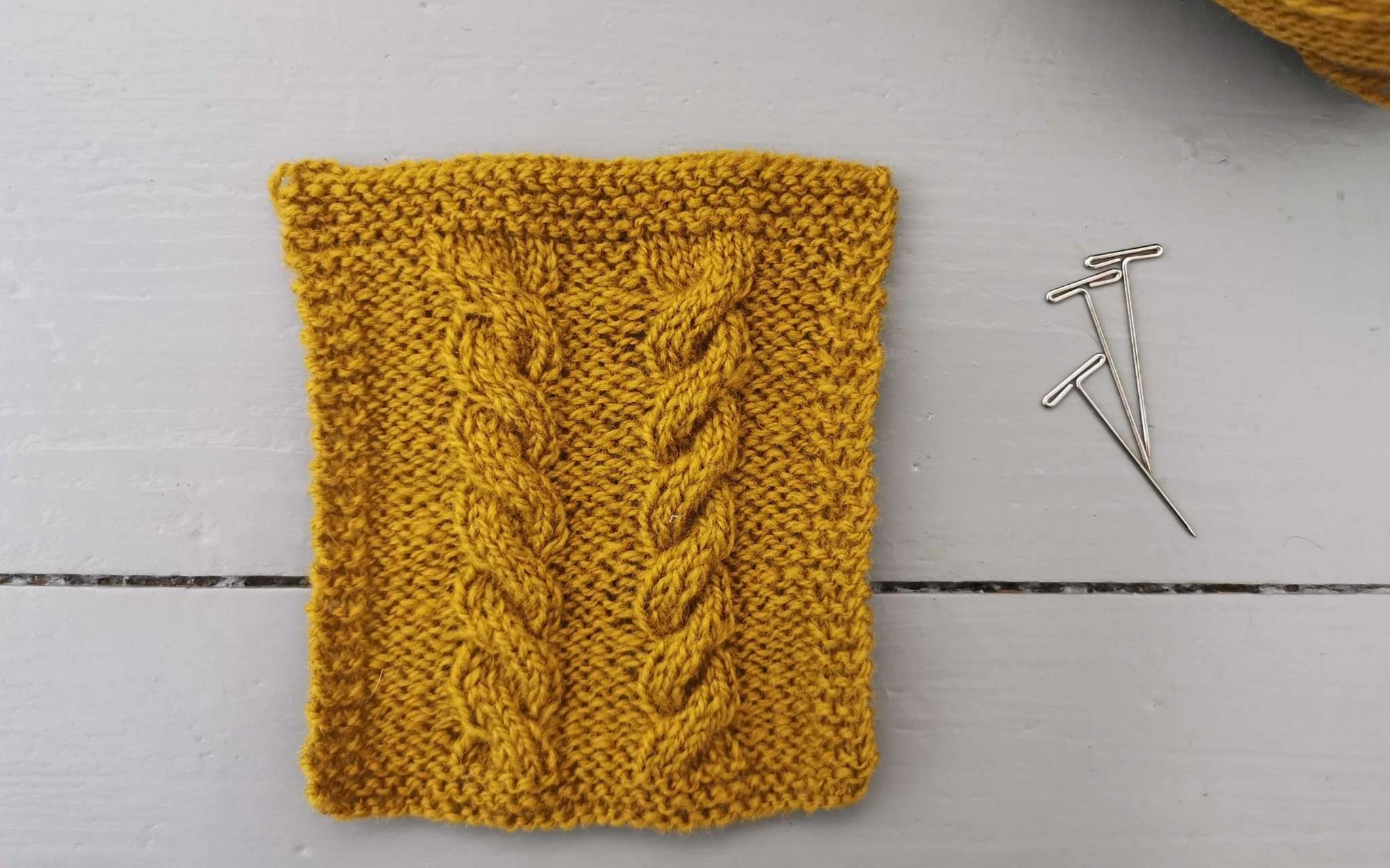 a golden yellow swatch with two cables running up the centre, photographed on a grey wooden background. Three t-pins are at the side and a glimpse of the ball of yarn used in the corner