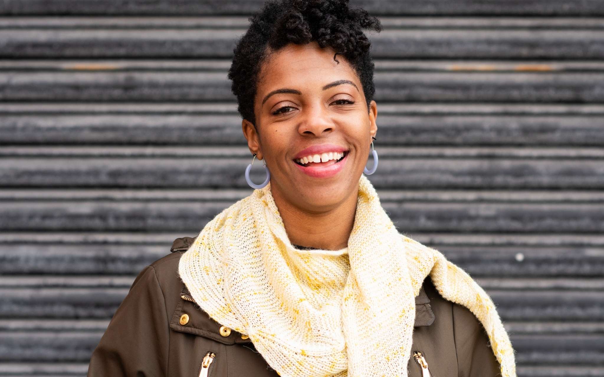 A young black woman with short hair and hoop earings stands in front of an iron door. She is wearing a pale lemon shawl with a brown jacket and is smiling.