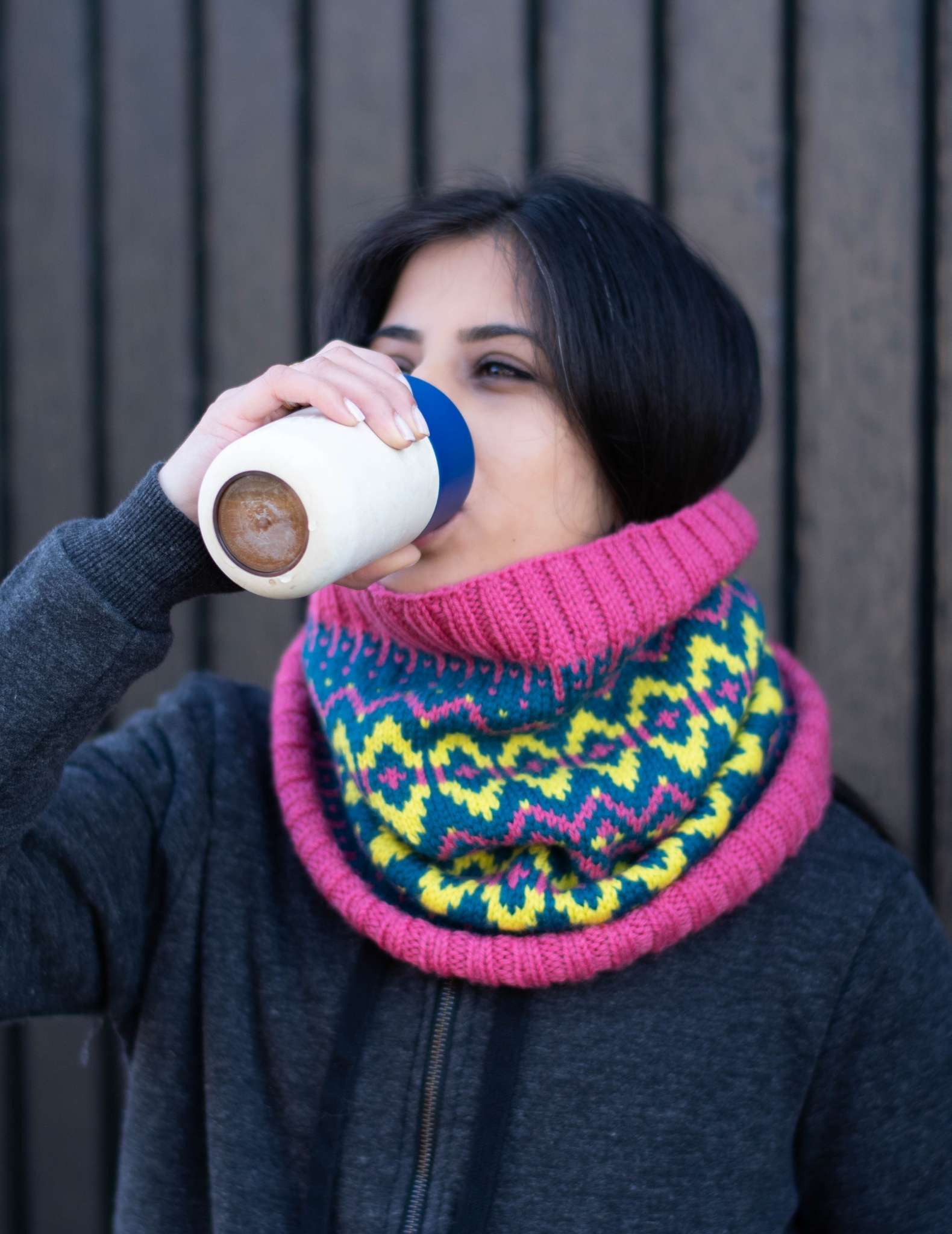 a woman with dark hair wears a pink, green and teal colourwork cowl, she is drinking from a coffee cup