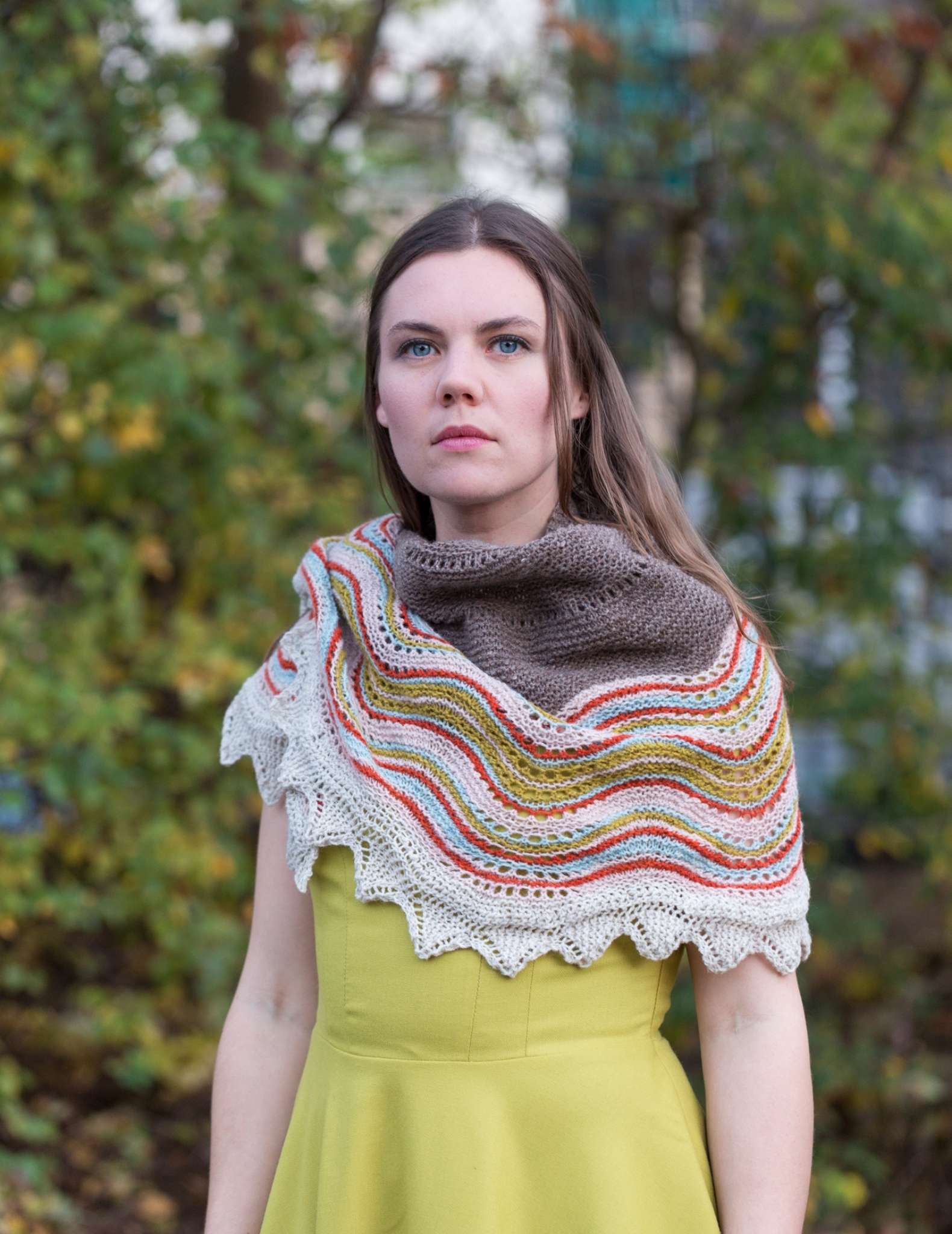 A white woman with long brown hair stands outdoors with blurred tress behind. She is wearing a large shawl with a striped edging and brown centre, wrapped around her neck with a yellow dress. She looks straight at the camera.