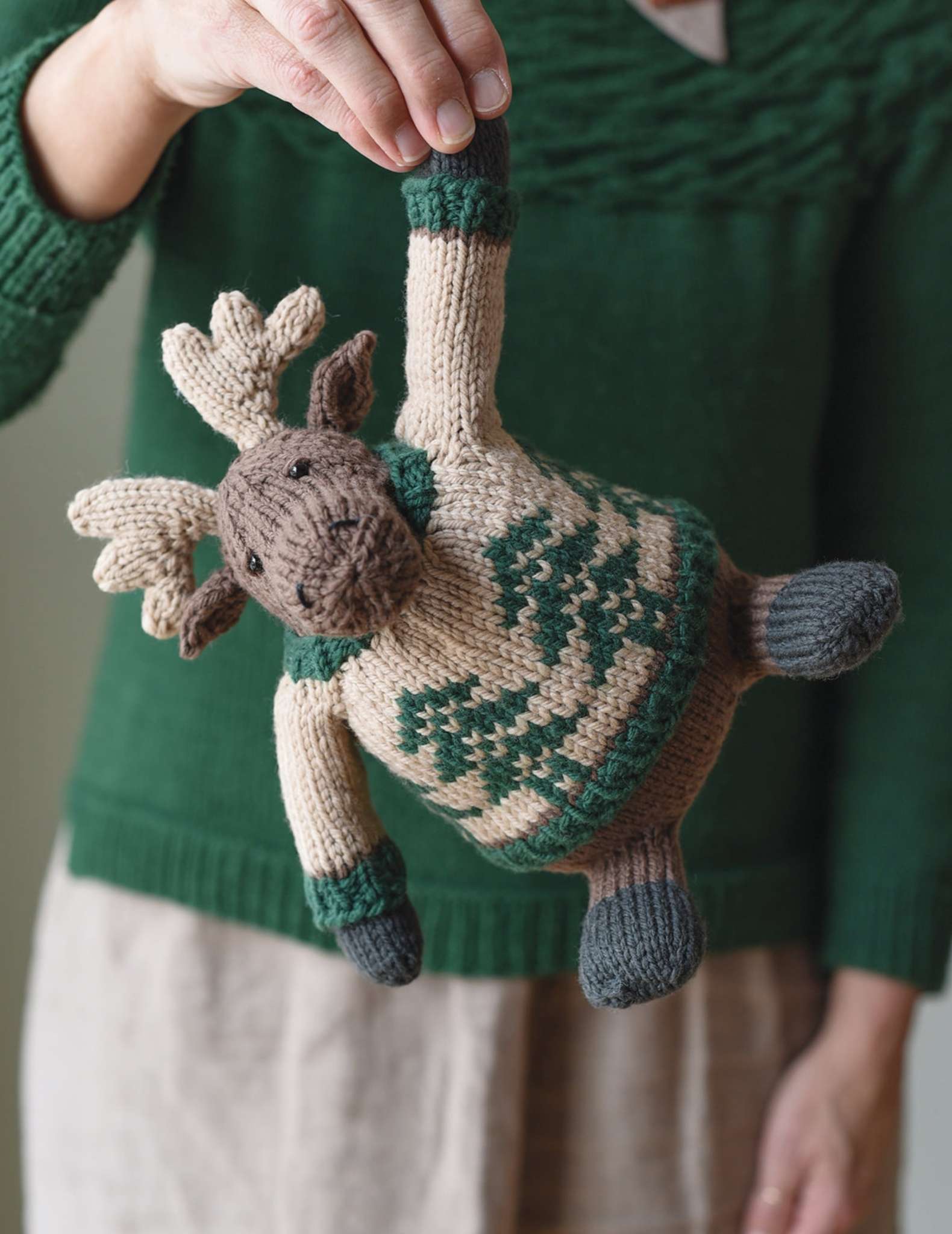 A knitted toy moose is hanging down in front of a model, being held by one arm. The moose wears a colourwork sweater with a tree motif. The model has white hands and is wearing a green sweater.