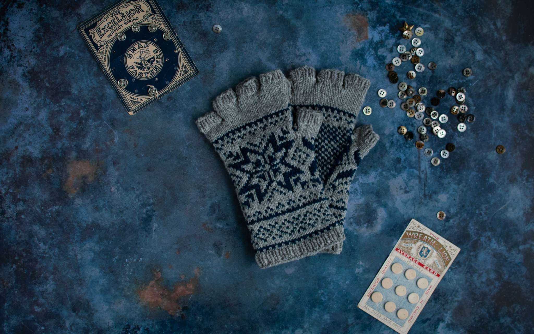 A pair navy and grey colourwork fingerless gloves lay flat on a moody blue background. Next to them are piles of blue and grey vintage buttons.