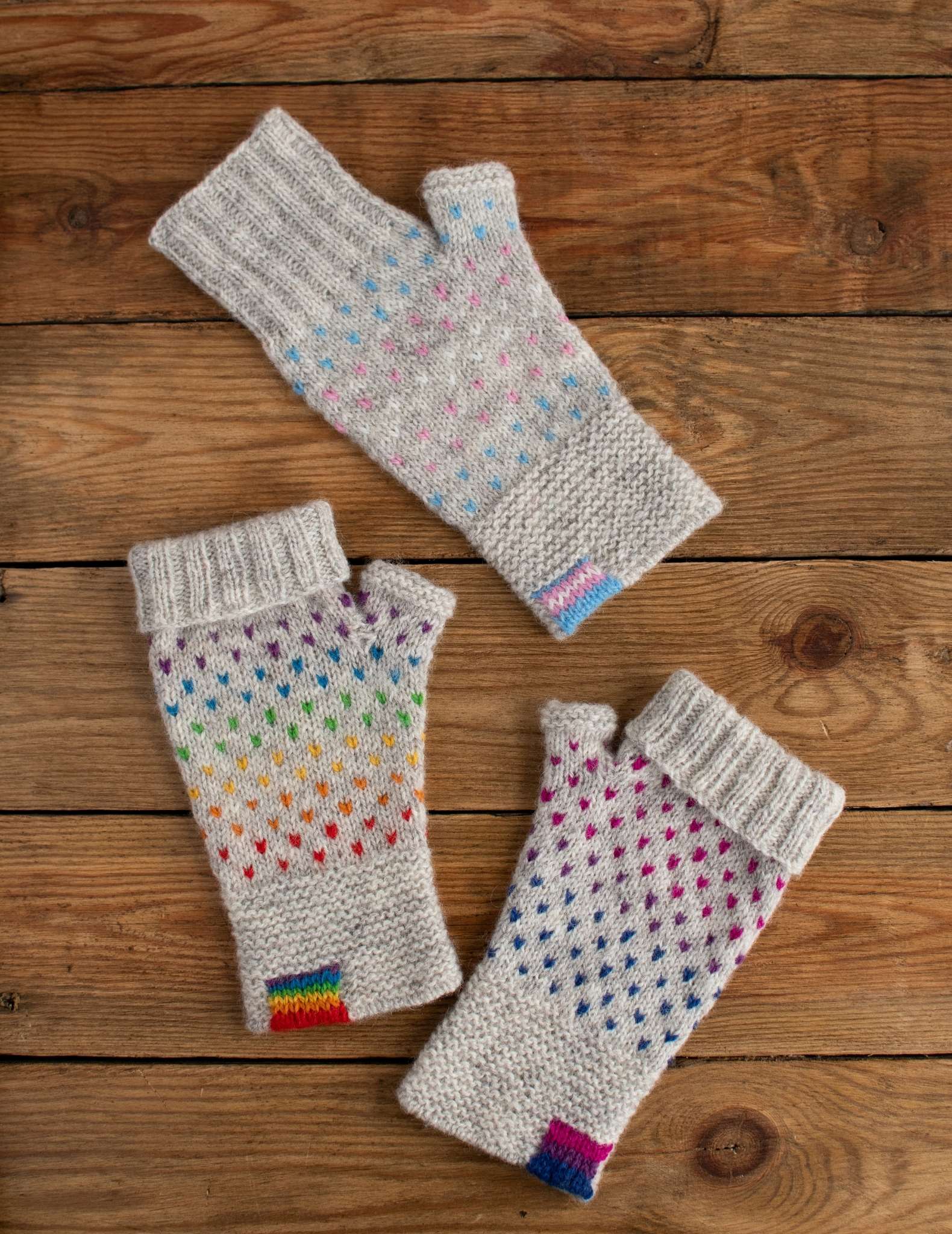 three individual fingerless mitts lay flat on a wooden surface. Each features a dot pattern on a grey background and a flag on the cuff in pride, bi, and transgender colours