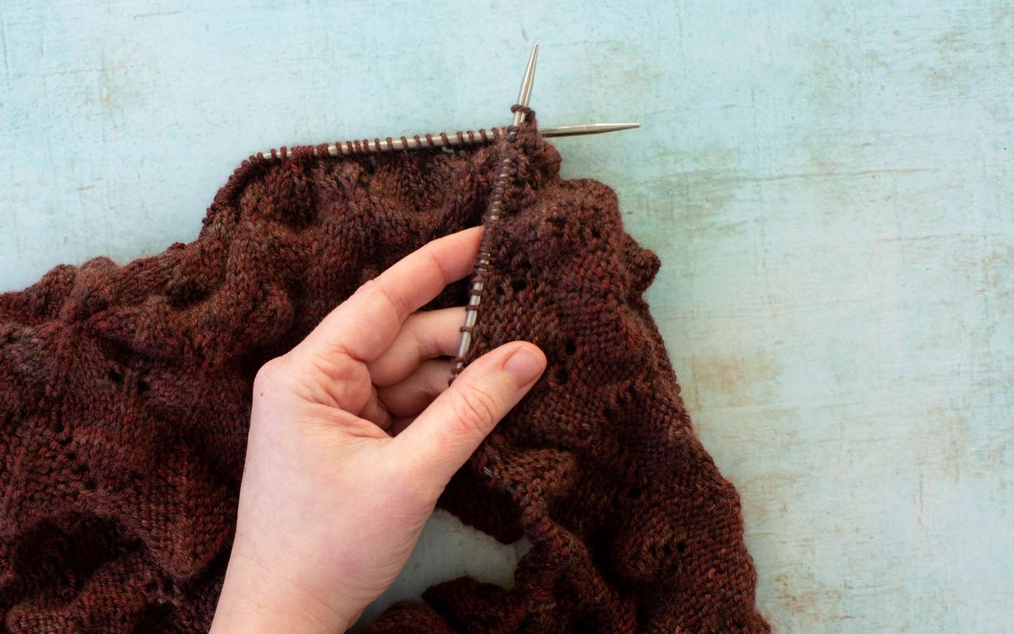 A hand holds up a piece of brown lace knitting, folded over to show both the right and wrong sides of the work.