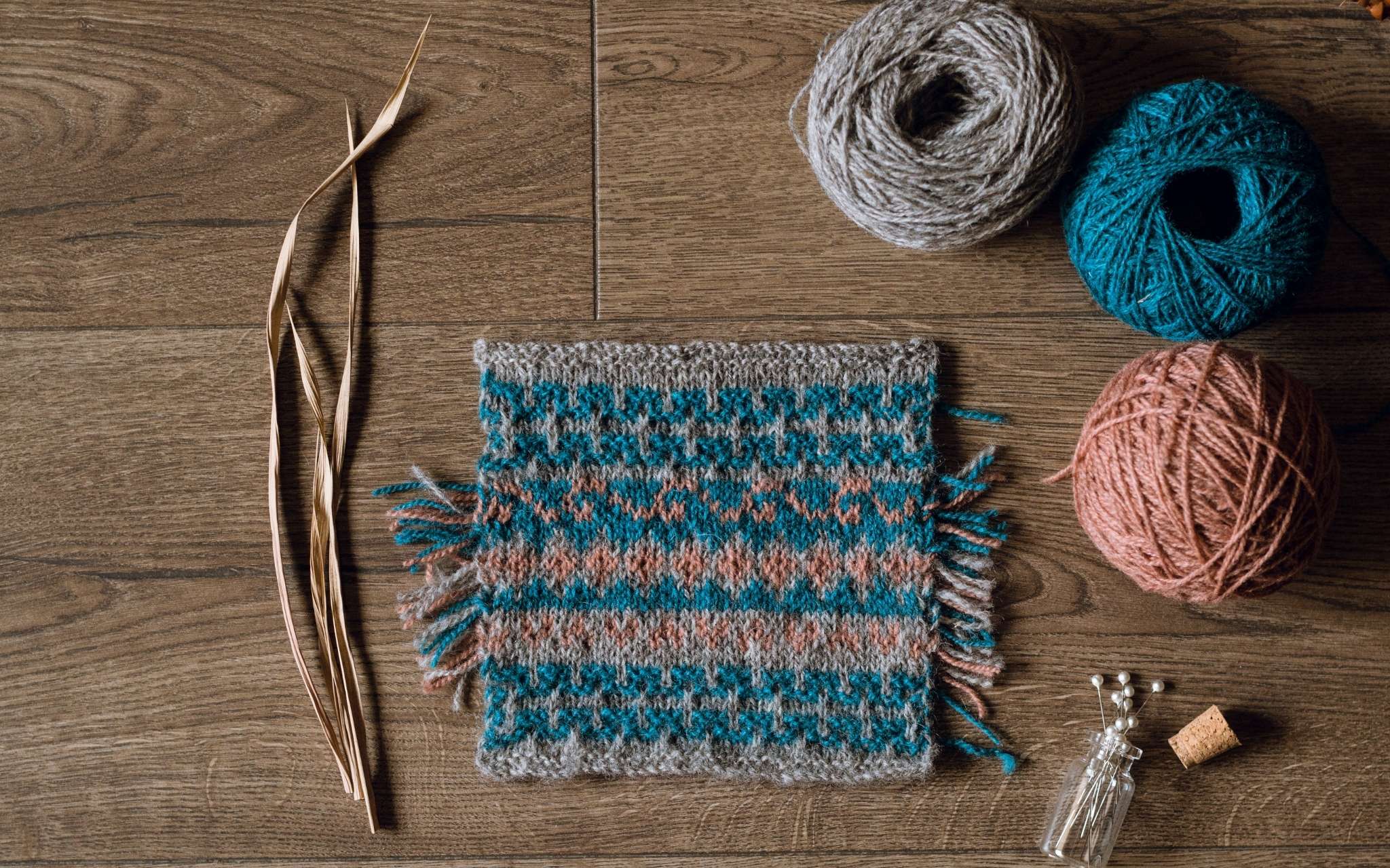 A swatch of colourwork knitting in dark blue, coral and grey laid flat on a wooden surface. There are some dried stems, scissors and a bottle of pints to the side.