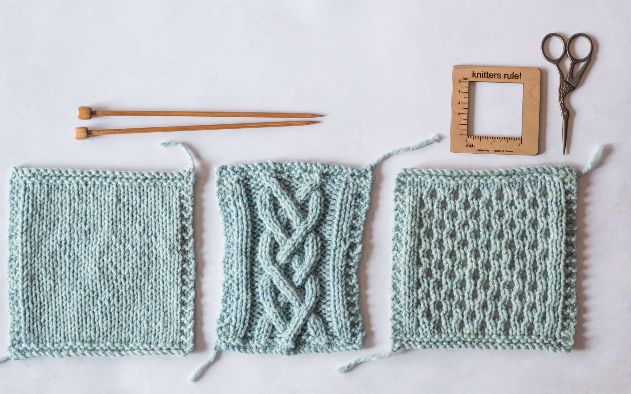 Three pale blue swatches lie next to each other, the first is in stocking stitch, the middle is in a cable pattern and the third shows a lace pattern. Above the swatches are a pair of straight wooden knitting needles, a square wooden measuring tool and a small pair of scissors.