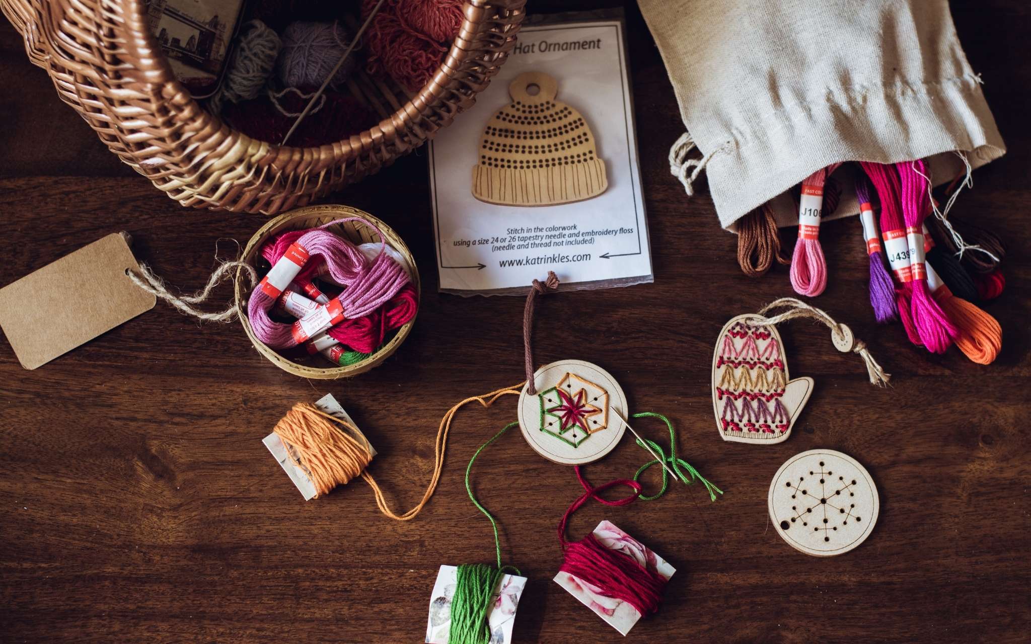 A selection of wooden hanging ornaments lie on a flat surface next to a wicker basket, and a pile of brightly coloured threads.