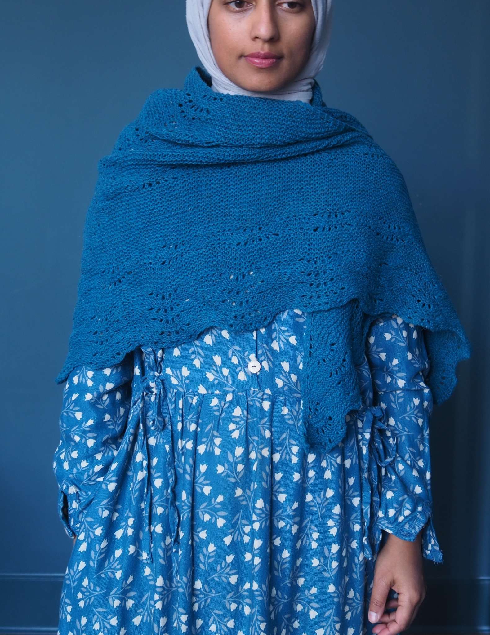 A brown woman in a hijab stands in front of a dark blue background. She is wearing a blue dress with small white floral print and a dark blue shawl which is wrapped around her chest and shoulders.