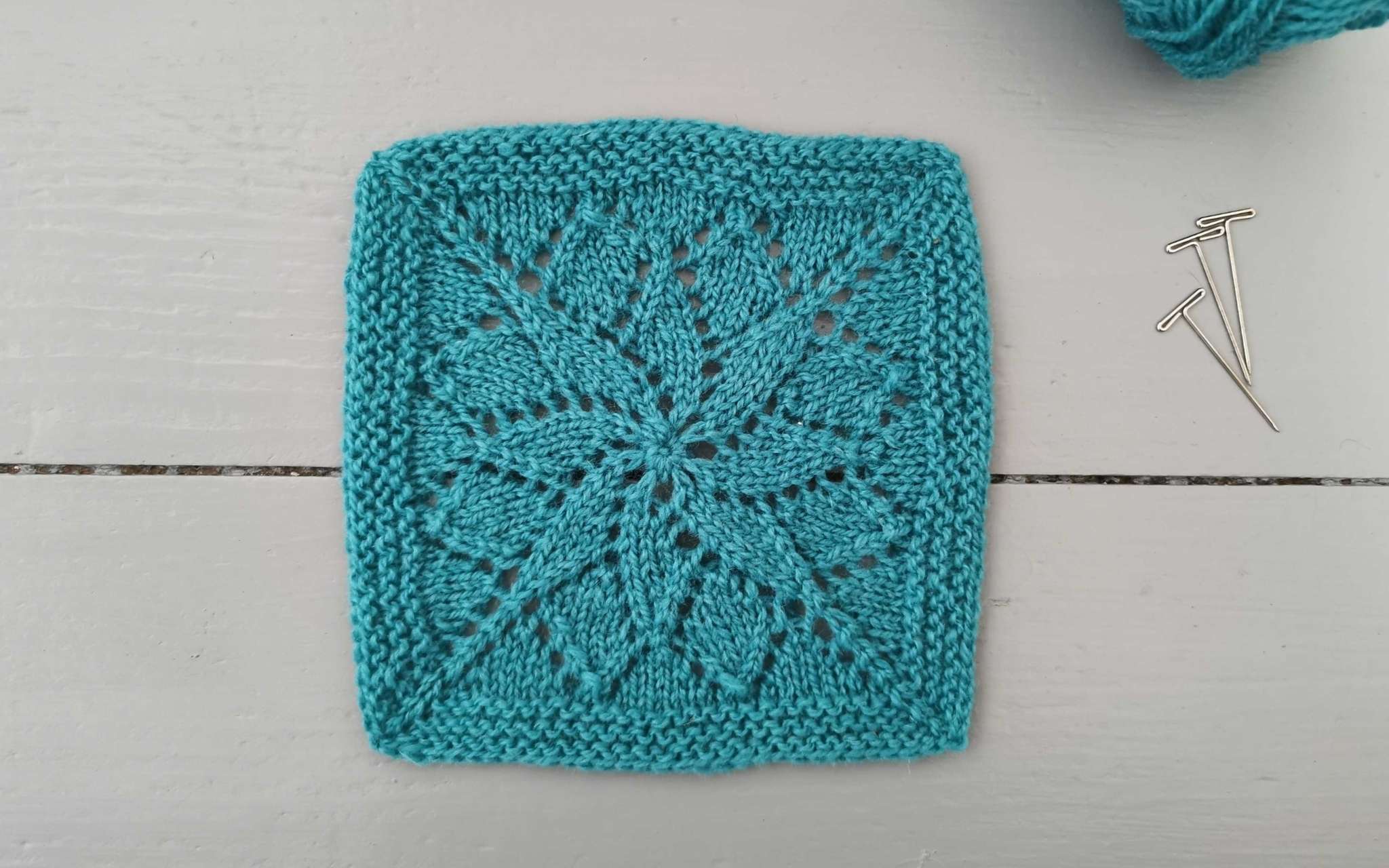 a square lace swatch of knitting, with three t-pins to the right and a glimpse of the ball of yarn used.