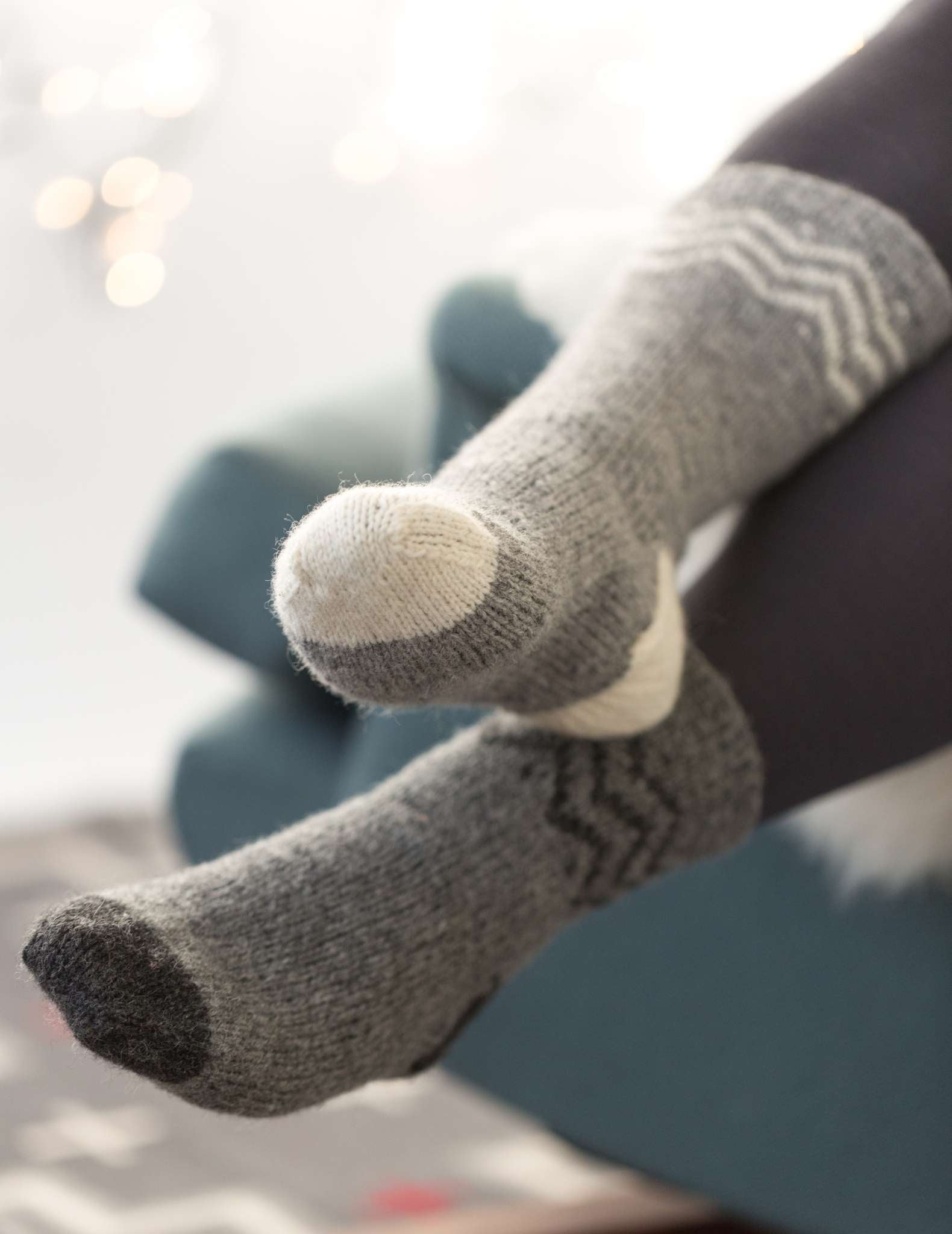 A model wears a pair of grey socks with colourwork around the cuff and contrast heels and toes. The contrast for one sock is white, and the other is dark grey.