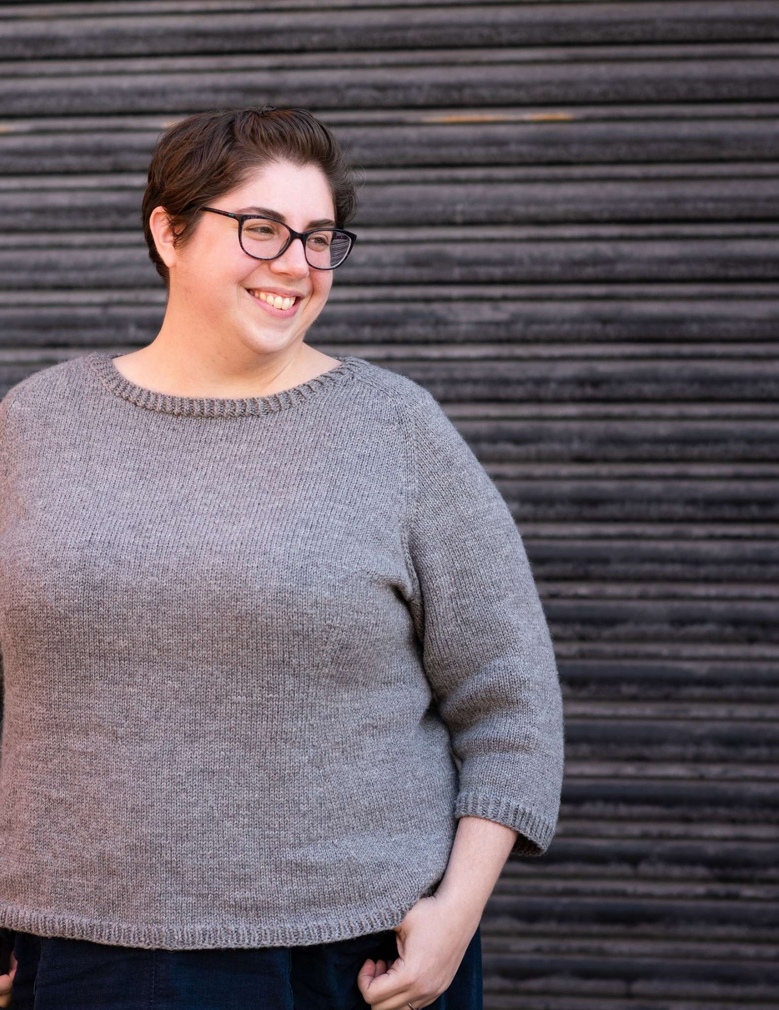 A white plus-size woman with dark hair and glasses wears a grey sweater, she is smiling and looking to the side with one hand resting on the top of a pocket