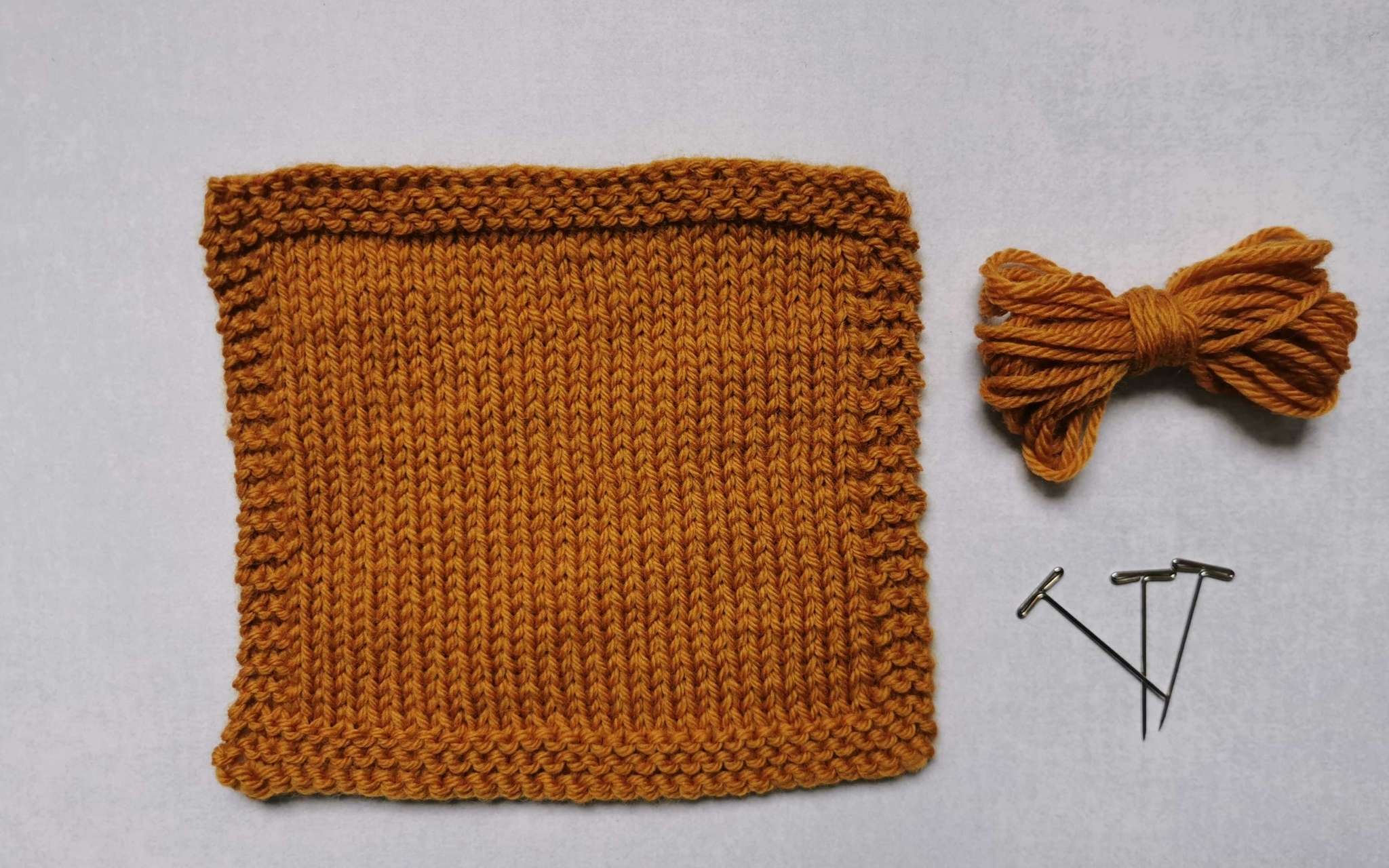 A square swatch of stocking stitch in golden yellow, with some bundled up spare yarn and t-pins laying beside it to the right.