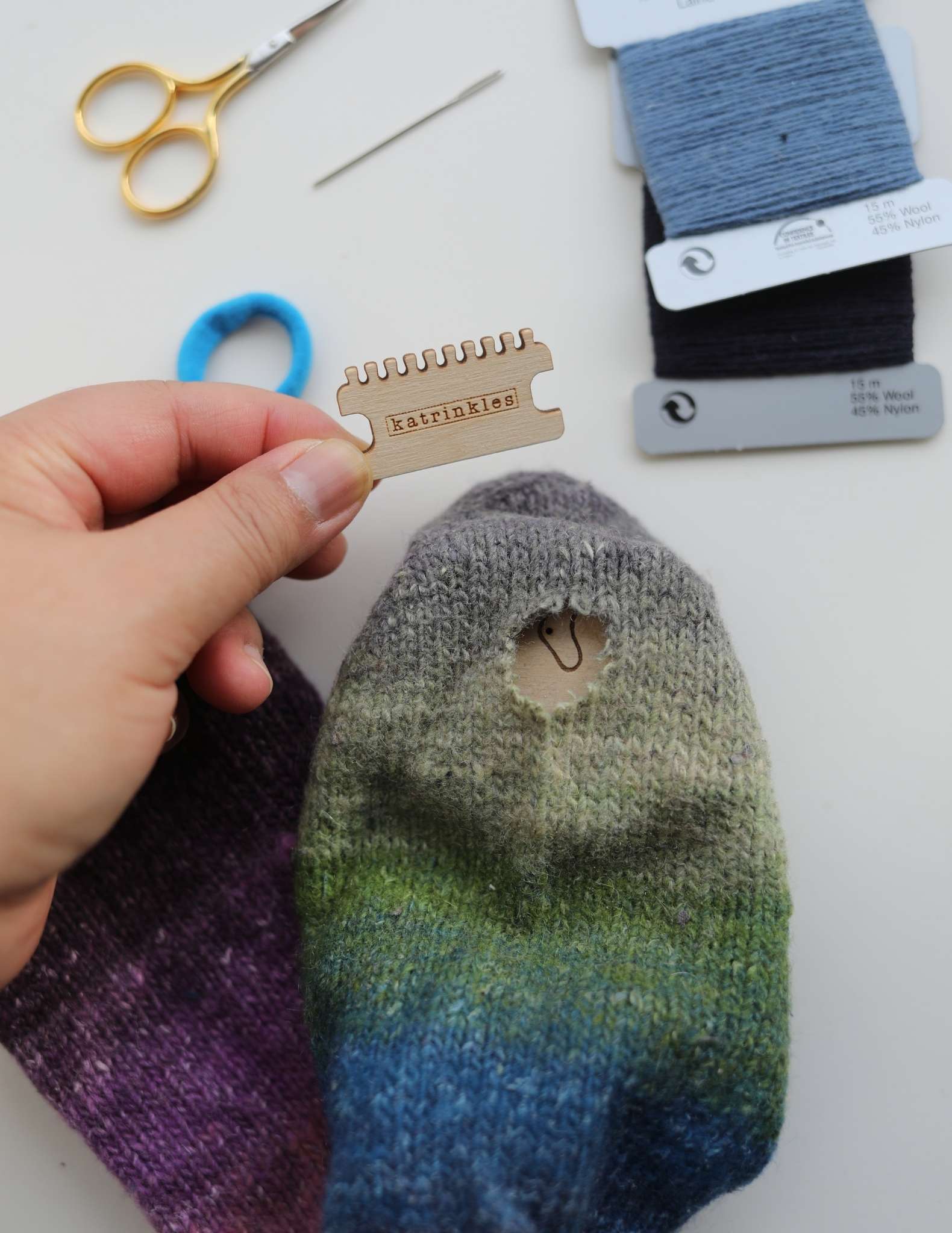 A sock showing a hole in the foot, and underneath the hole is a wooden darning loom, with the wood visible through the hole. A hand to the left holds a small wooden heddle.