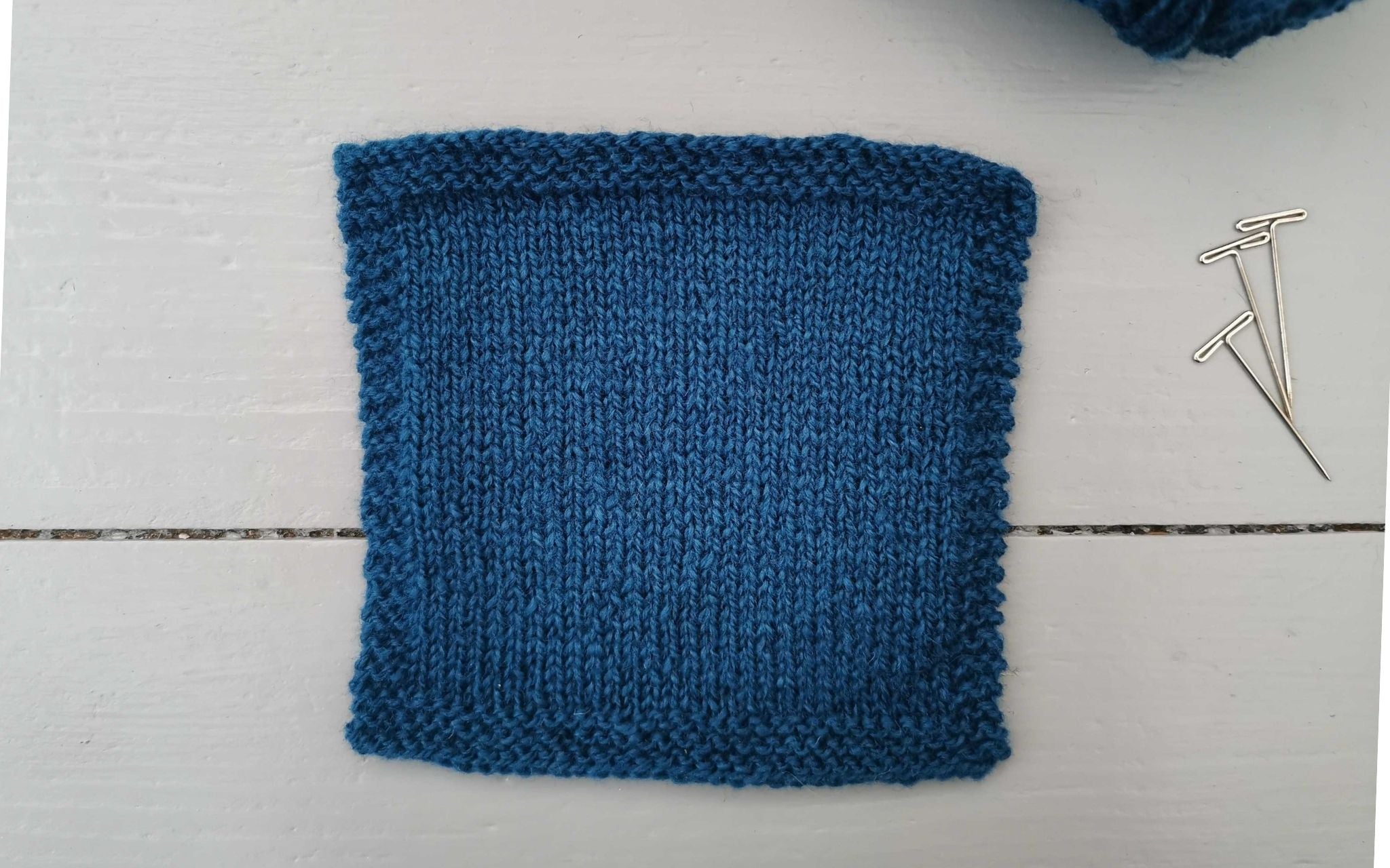 a square navy swatch of knitted fabric, with a tiny glimpse at the ball of yarn used in the corner and three t-pins at the side