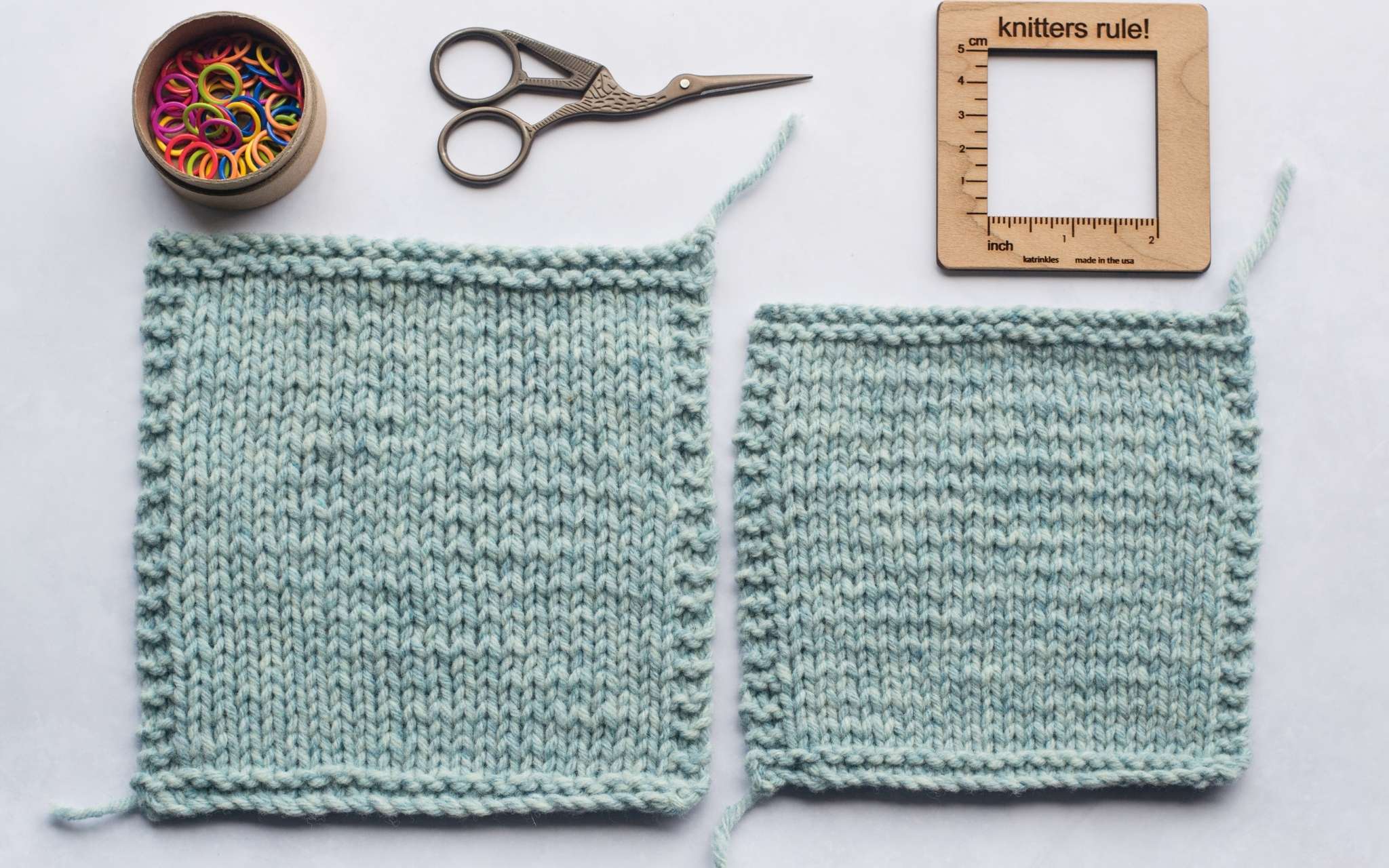 Two pale blue swatches lie on a flat surface. The left swatch is bigger and knit at a looser tension, the right swatch is smaller and a denser fabric. Above the swatches are a pot of brightly coloured stitch markers, a small pair of scissors and a wooden square measuring tool.