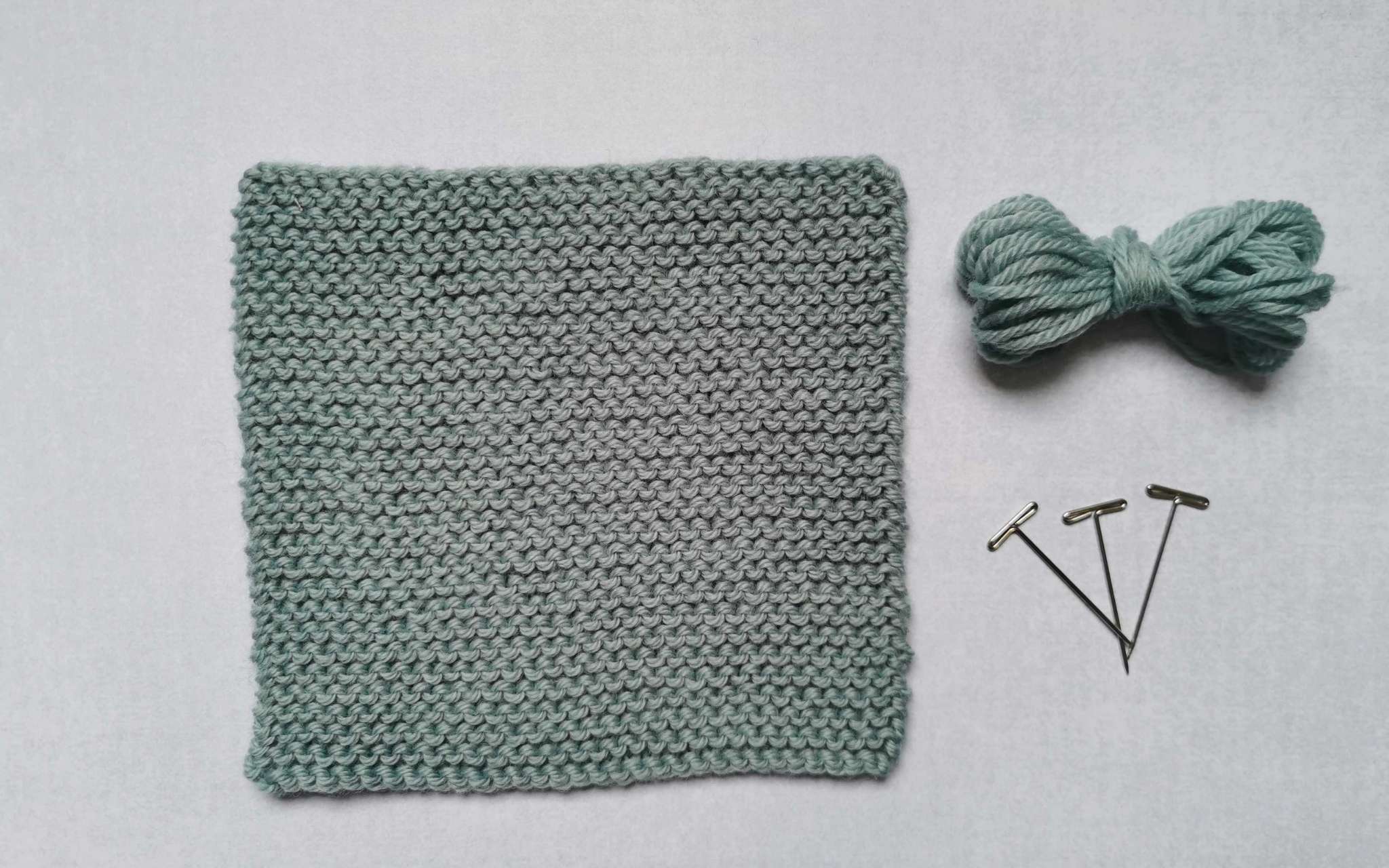 A square swatch of stocking stitch in pale blue, with some bundled up spare yarn and t-pins laying beside it to the right.