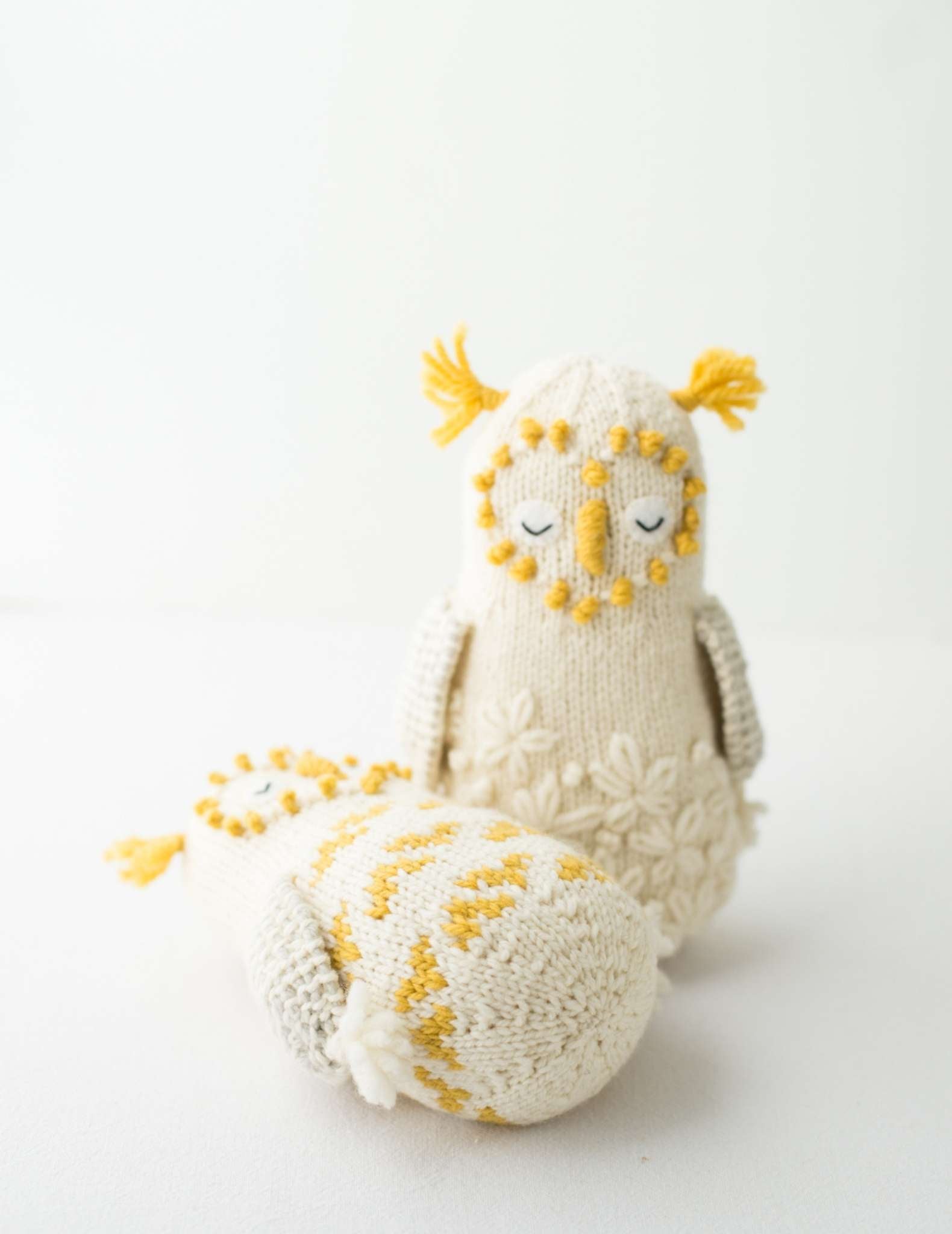 Two white knitted owls, with yellow details on the ears and face. One stands up straight at the camera, while the other lays down on its back beside the other.