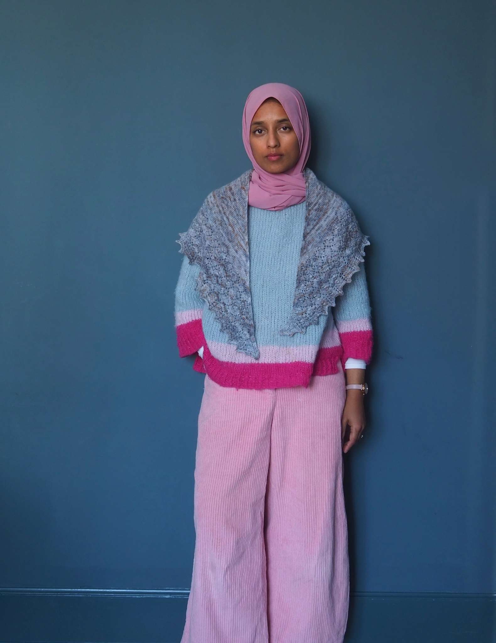 A brown woman in a pink hijab stands in front of a blue background looking straight into the camera. She is wearing a pale pastel jumper in blues and pink, pale pink trousers and a grey blue shawl around her neck with the two points hanging down in front.