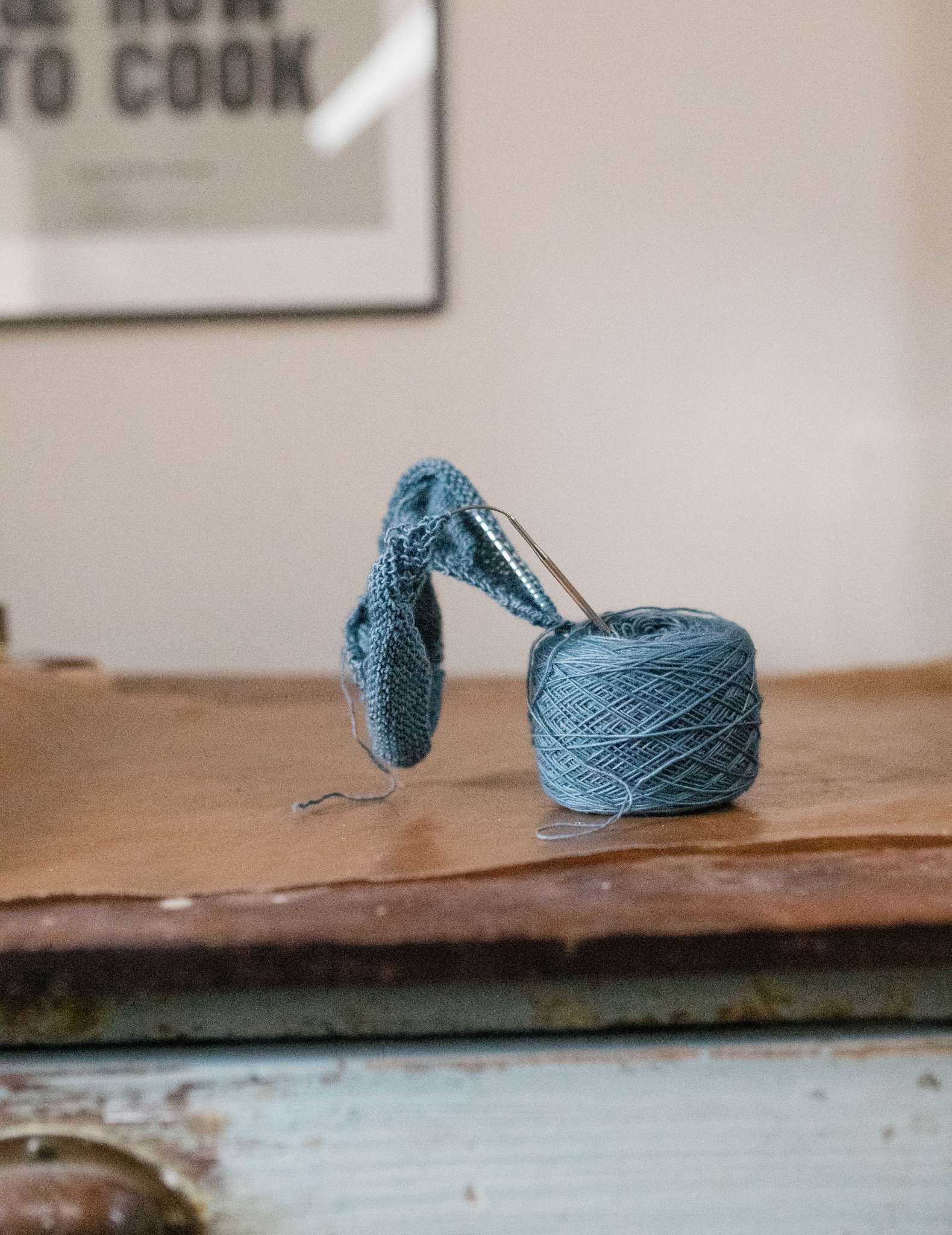 A ball of blue yarn with a project on the needles sticking out of the top of it, sitting on a wooden surface in front of a wall with an art print.