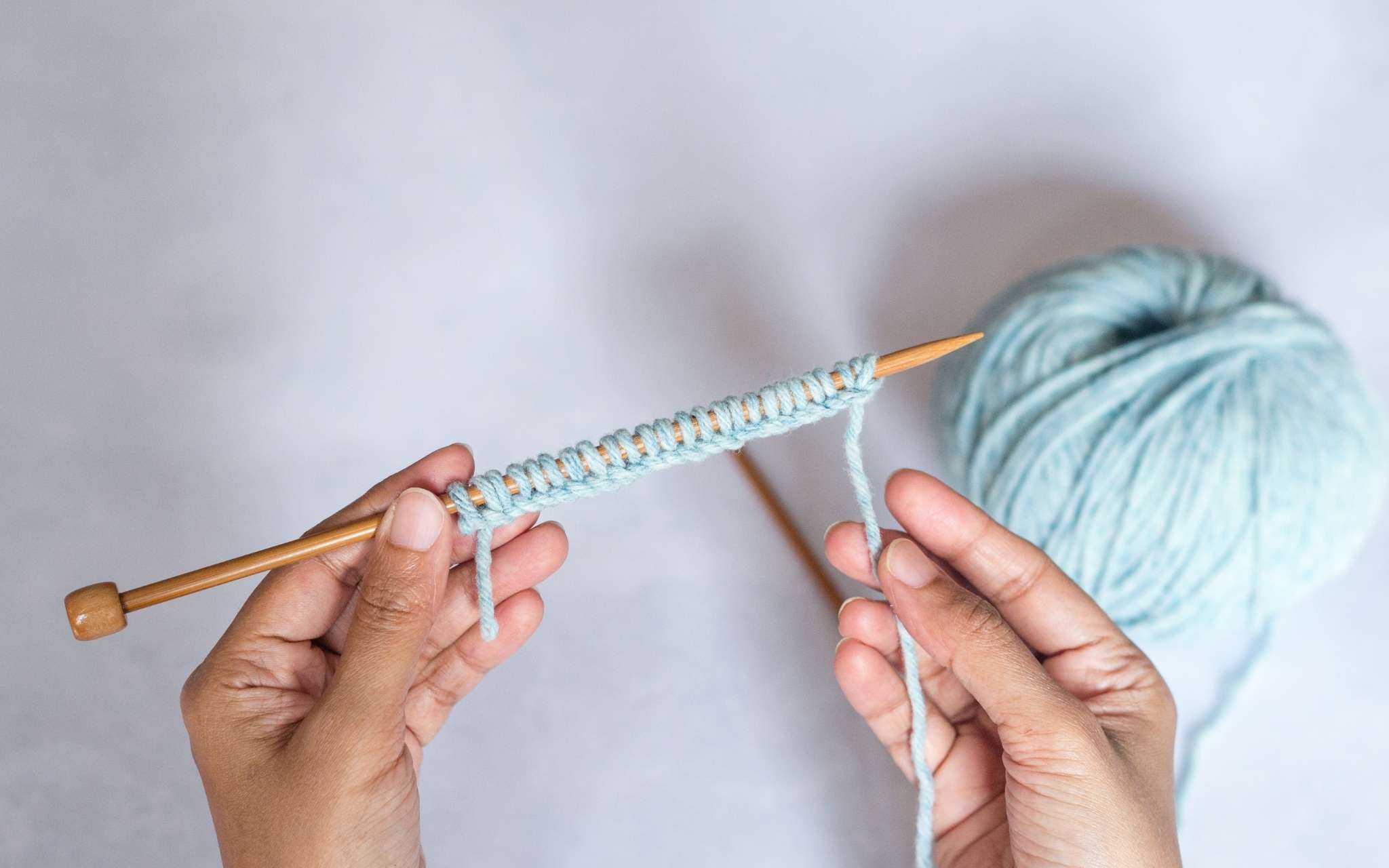 A woman of colour holds a cast on row of stitches on a straight knitting needle. The stitches are in pale blue yarn and the ball of yarn lies to the right of the image.
