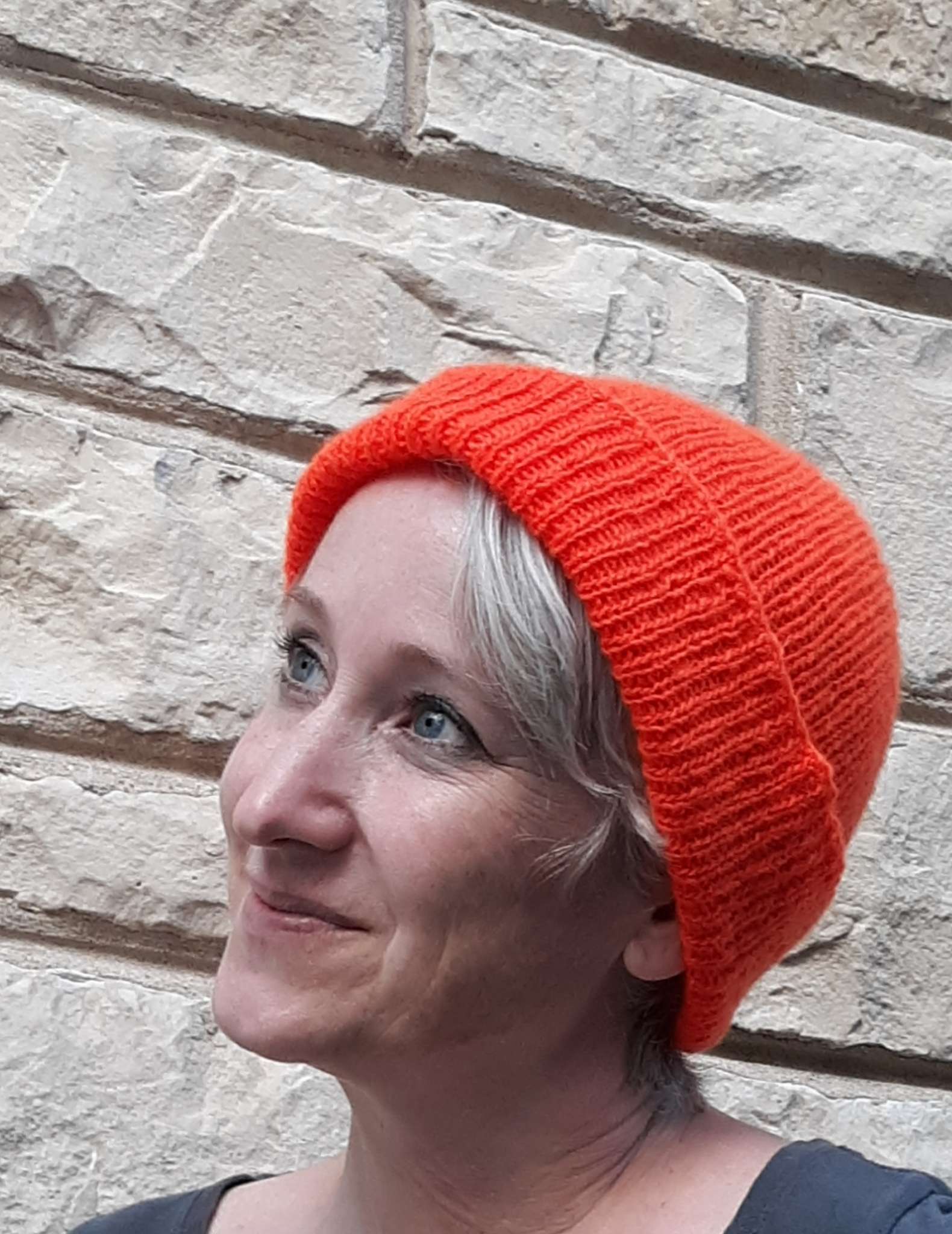A smiling person with short grey hair is pictured wearing a bright orange ribbed hat with the brim turned up in front of a white brick wall. 